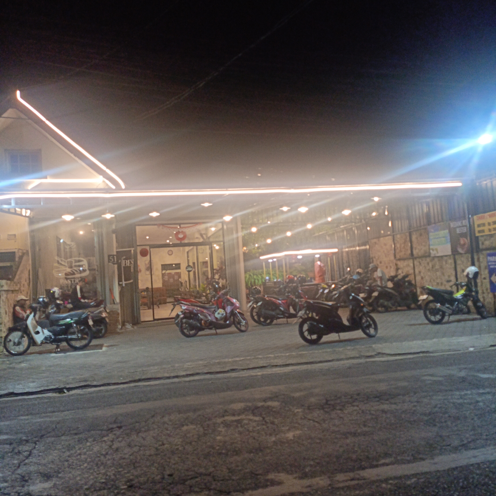 The image depicts the exterior of a building at night, likely a cafe or restaurant, with a bright, illuminated facade. The building features large glass windows and doors, revealing an interior with tables and chairs, and decorative lights. In front of the building, a row of parked motorcycles is visible, suggesting a popular spot for patrons arriving on two-wheelers. The ground in front of the building is paved with asphalt, showing some wear and tear, and there are streetlights or decorative lights illuminating the scene. The overall impression is a lively urban setting at night. A sign on the building reads "51 BES" and another sign in the background appears to be in Indonesian. Based on the context provided by the user, this scene is located in the city of Salatiga, Indonesia.