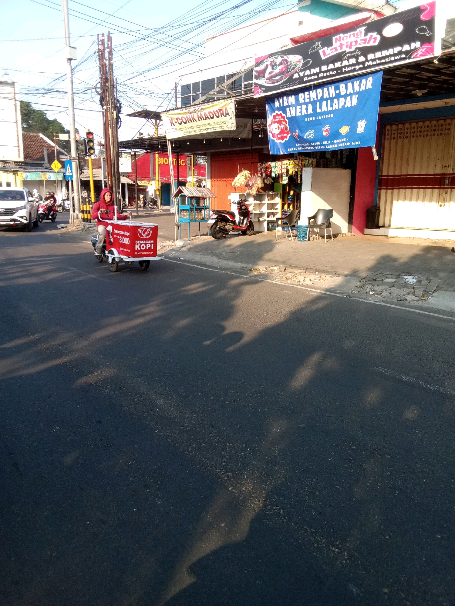 A man wearing a red hooded sweatshirt rides a three-wheeled scooter with a red refrigerated box attached to the back, in Sumber Lor, Indonesia. The box has white text that reads "SEMACAM KOPI" and "3.000" with a coffee cup icon. The scooter is on the right side of the road. To the left of the scooter, a white car and two motorcycles are visible. Overhead, a complex network of wires and utility poles crisscrosses the sky. Buildings line the street, featuring various signs for businesses. One sign, with a pink and black design, advertises "Neng Latipah" and "AYAM BAKAR & REMPAH" (grilled chicken and spices). Another, a blue banner, reads "AYAM REMPAH-BAKAR ANEKA LALAPAN NENG LATIPAN" (grilled chicken and spices, various side dishes). A smaller sign reads "K-CONK MADURA". The street is paved and shows signs of wear, with some broken sections and debris. Sunlight casts long shadows across the road.
