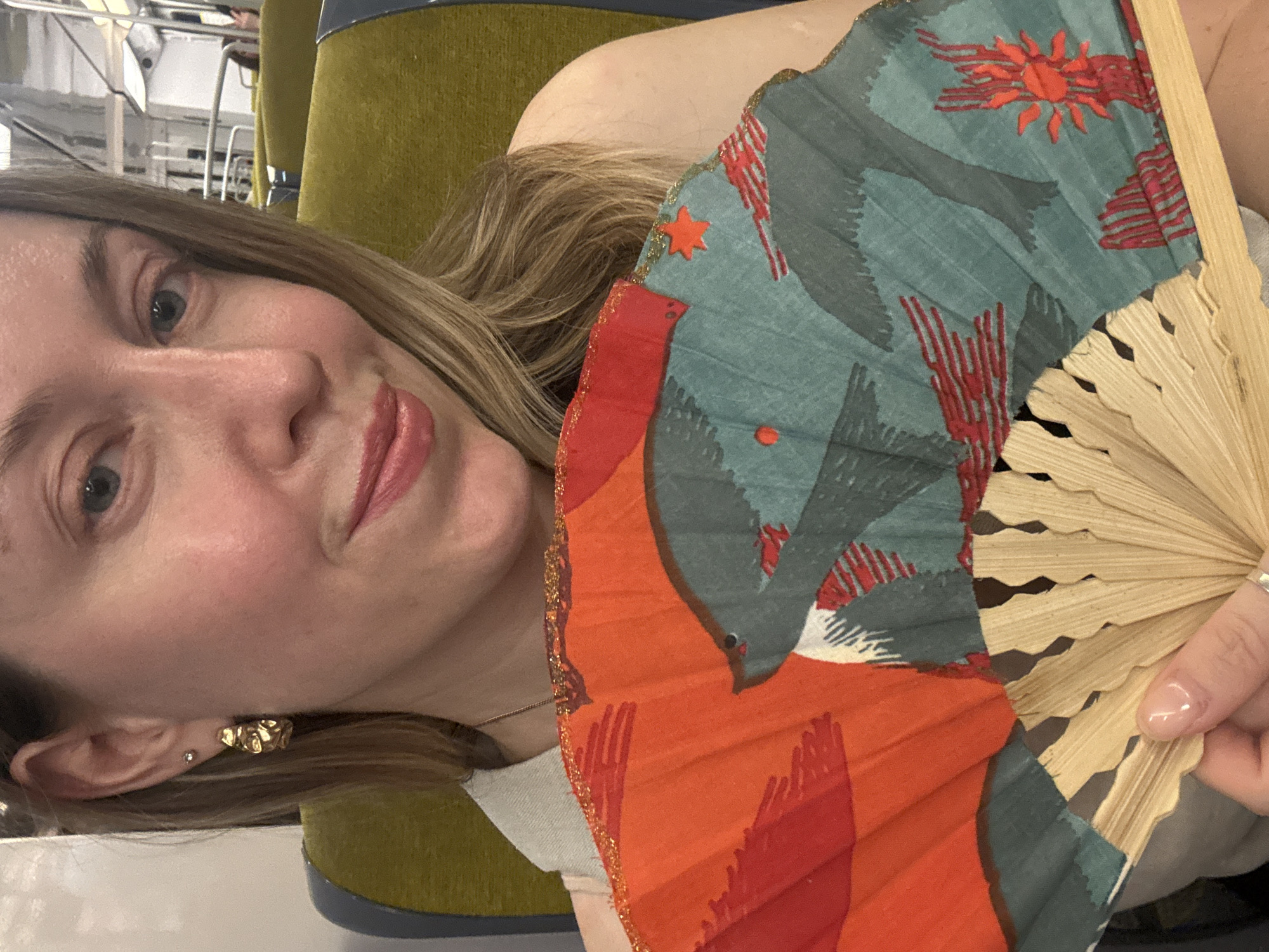 The media file is a selfie taken by a young woman in Gentilly, France. She is holding an open folding fan with a colorful pattern of birds and abstract shapes in shades of teal, red, and orange. The fan has a wooden handle. The woman has blonde hair and is wearing makeup, including lipstick and subtle eyeshadow. She has a small stud earring in her left ear and a more ornate gold earring in her right ear. She is wearing a light-colored top. The background is blurry but appears to be an indoor setting with hints of a muted green chair and some metallic structures that might be part of a public transport or building interior. The lighting suggests it could be daytime. The woman's expression is relaxed and she appears to be enjoying herself, as indicated by her slight smile and the way she is holding the fan.