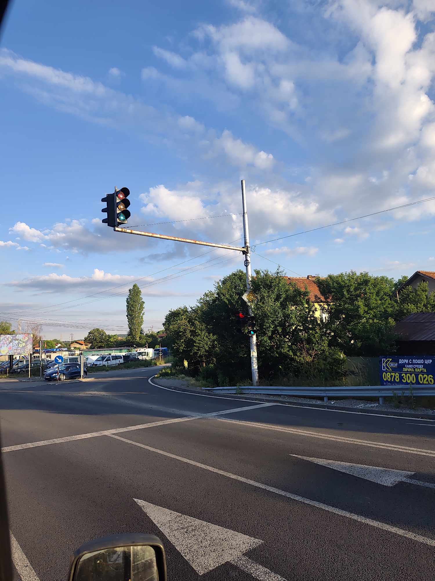 The image depicts an outdoor scene at an intersection in Dragichevo, Bulgaria, on a partly cloudy day. A traffic light, with red and yellow lights illuminated, is positioned on an overhead arm extending across the intersection. The road surface is asphalt, with white lane markings, including an arrow indicating a turn or lane. In the background, there are trees, buildings with red-tiled roofs, and a tall, slender tree. Several vehicles, including cars and vans, are visible parked or moving near the intersection. A metal guardrail runs along the side of the road, and a billboard with text and a phone number is mounted on it. The perspective suggests the photo was taken from inside a vehicle, with a side mirror visible in the lower right corner.