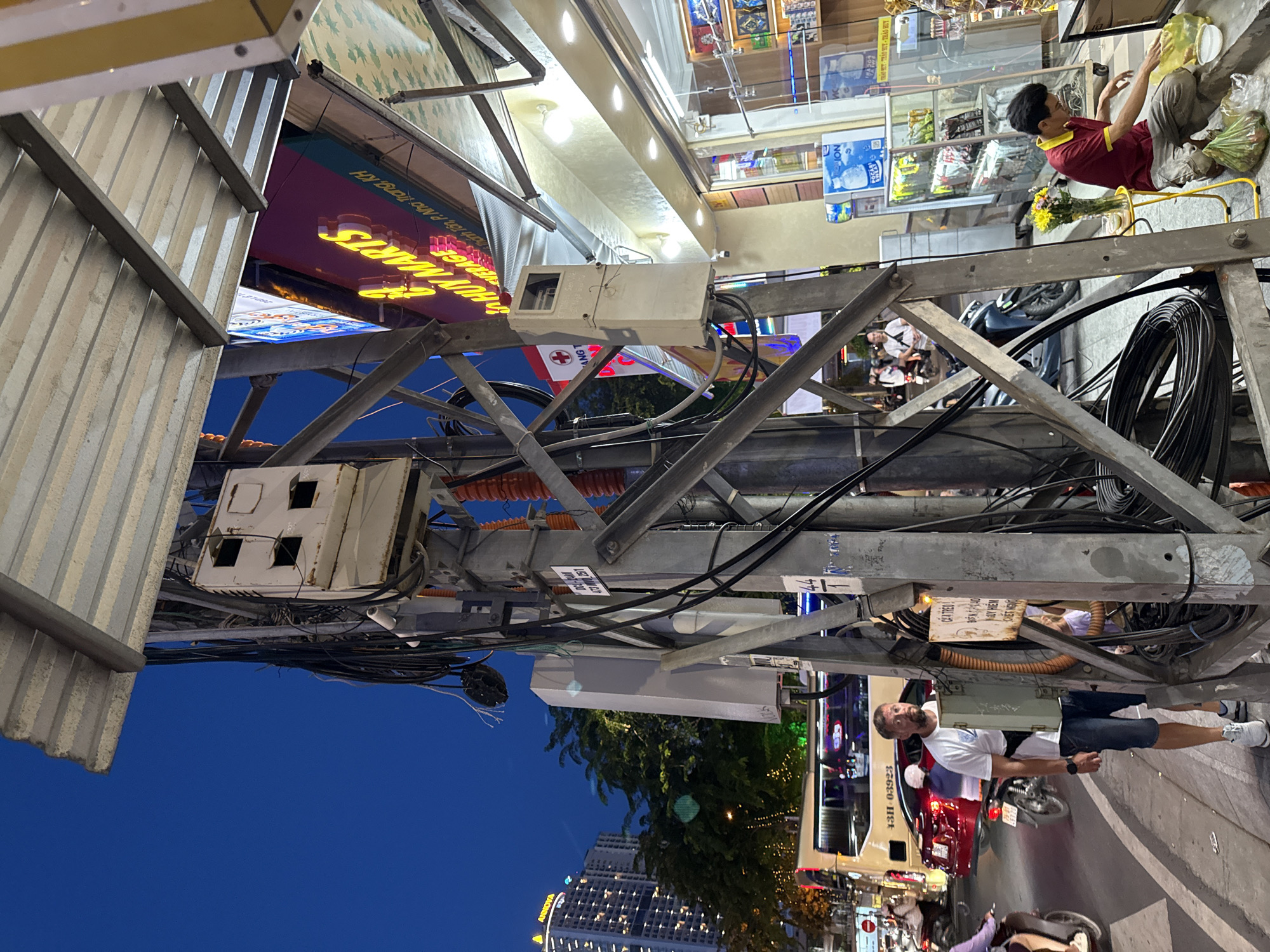 This is a street-level photograph taken in Nha Trang, Vietnam, likely at dusk or early evening, given the deep blue sky and illuminated storefronts. The image is dominated by a complex array of electrical poles, wires, and junction boxes, indicative of urban infrastructure. A large metal pole with numerous cables and several gray, box-like enclosures is centrally positioned. One enclosure has multiple openings, suggesting it houses electrical components. Another, longer gray box is mounted lower on the pole.
To the left, a street scene unfolds. A white tour bus with the license plate "43H-03923" is visible, along with a red car and several motorcycles. A man wearing a white t-shirt, denim shorts, and white sneakers stands near the pole, looking towards the right. He has a black fanny pack slung across his body. Further down the street, other pedestrians and vehicles are present.
To the right of the main pole, a storefront with bright neon signage that reads "HUY MARTS" is illuminated. The store appears to be a convenience store or market, with shelves visible through the glass facade displaying various products. A man in a red shirt and beige pants is kneeling in front of the store, interacting with plastic bags and a small yellow stool with a vase of flowers on it. The overall environment suggests a bustling urban commercial area. The lighting is a mix of natural twilight and artificial light from the storefronts and streetlights, creating varied shadows and highlights.