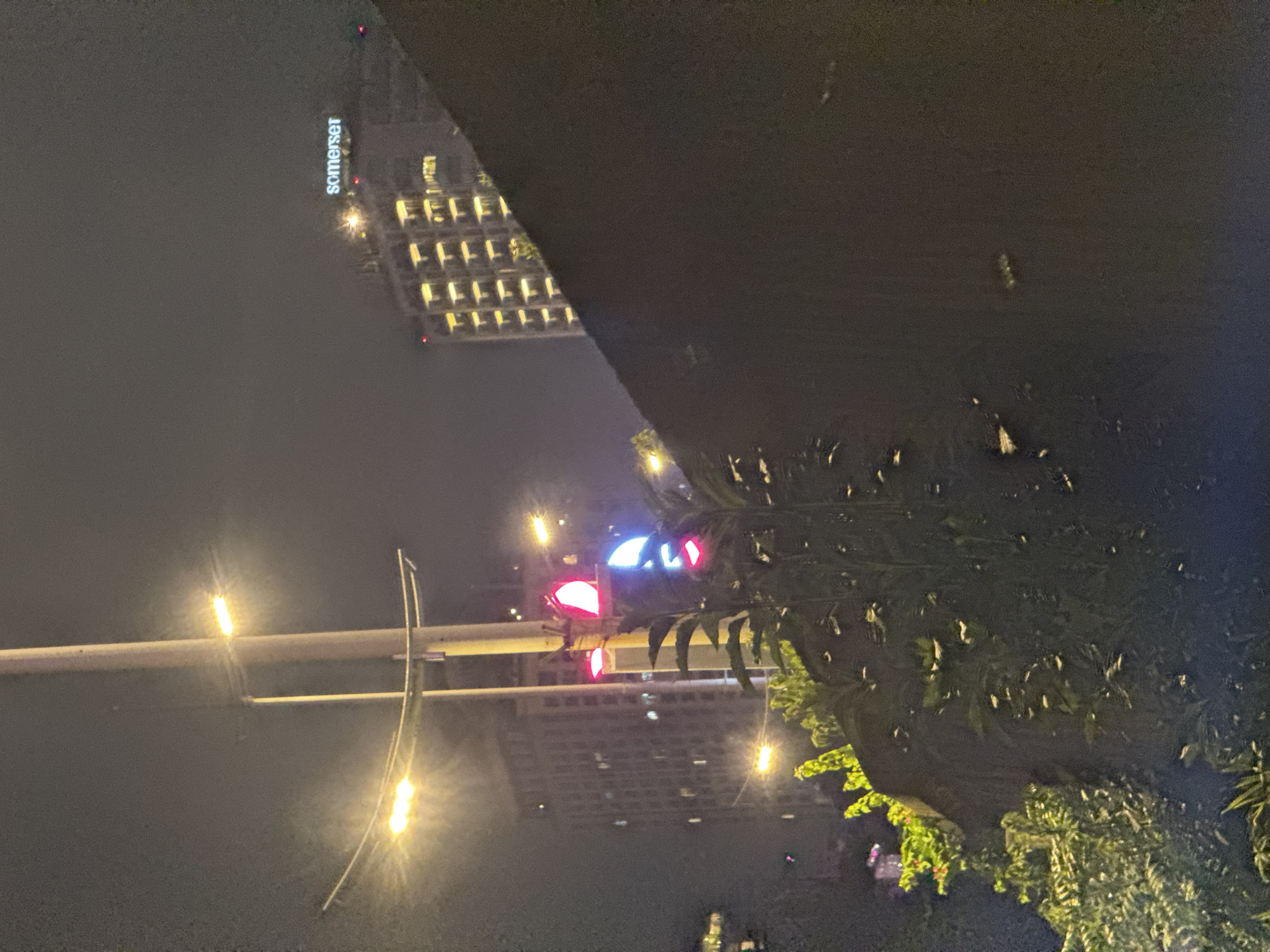 The image depicts a nighttime scene in Hanoi, Vietnam, likely near a building with "somerset" signage visible. The perspective is from a street-level view, looking upwards towards a dark, angular structure that dominates the upper portion of the frame. Below this structure, the lights of a building with a grid of illuminated windows are visible.
In the foreground, a traffic light shows red lights illuminated, indicating a stop. A street lamp with a bright yellow glow is also prominent on a pole. Trees with dense green foliage are positioned to the right, with some lights reflecting on their leaves, suggesting they are illuminated by nearby artificial light sources. The overall atmosphere is dark and hazy, typical of a city at night, with a slight blur suggesting motion or a low-light exposure. The image seems to be captured from a moving vehicle or handheld camera, as indicated by the slight tilt and lack of sharp focus on some elements.
