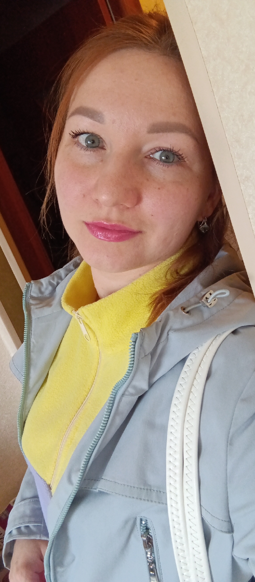 A woman with red hair and light blue eyes is wearing a yellow fleece jacket under a light blue jacket. She has freckles on her face and is wearing pink lipstick. She is standing indoors, and there is a door frame in the background. The location is Unknown, Unknown.