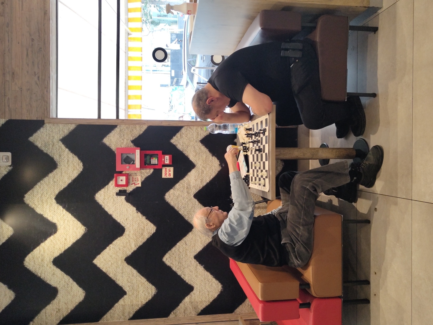Two men are engaged in a chess game in a public indoor space. An older man sits on a red and brown cushioned bench on the left side, his arm raised as he contemplates a move. Across from him, a younger man, dressed in black, leans over the chessboard. The chessboard is set on a table with wooden legs. A water bottle is visible next to the chessboard.
The background features a wall with a black and white zigzag pattern. Various red objects, resembling emergency equipment or decorative elements, are mounted on the wall. Behind the younger man is a window with horizontal yellow and white stripes. The floor is light-colored tile.
There is no immediate evidence to confirm that the scene is in Tel-Aviv, Israel, so that piece of information is omitted.