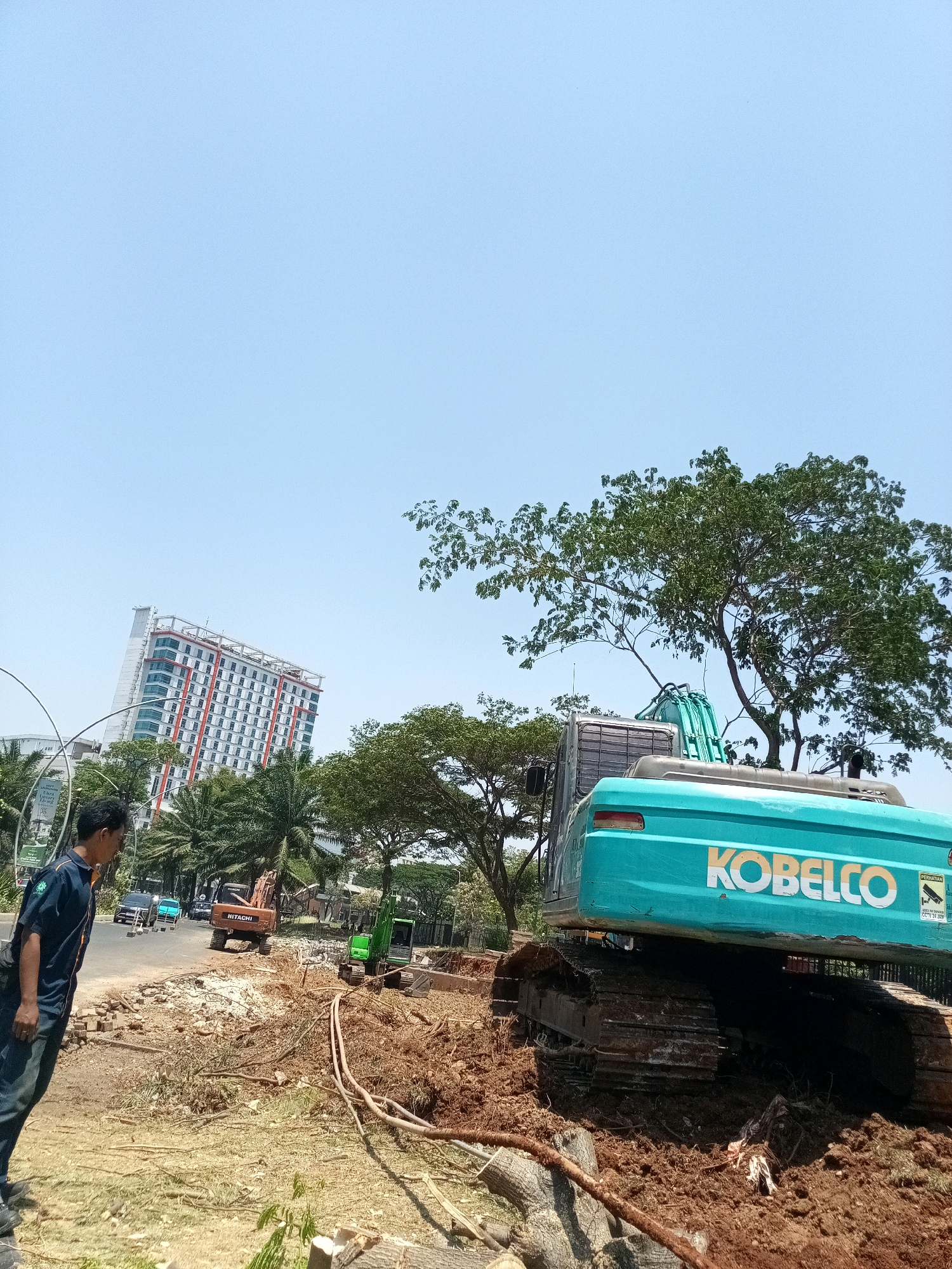 The image depicts a construction site in Bekasi, Indonesia, under a clear blue sky. A large turquoise Kobelco excavator is prominent in the foreground, its tracks deeply embedded in muddy soil. To the left, a man in a dark blue uniform with an embroidered logo on the left chest and a black bag slung across his body stands on a dirt patch, looking towards the construction activity. Behind him, a street with a few cars is visible. In the middle ground, a smaller green excavator and a reddish-brown Hitachi excavator are also at work. Further in the background, a tall, modern building with a white facade and red accents stands out amongst palm trees and other greenery. Debris and uprooted trees are scattered across the site, indicating ongoing land clearing or excavation.