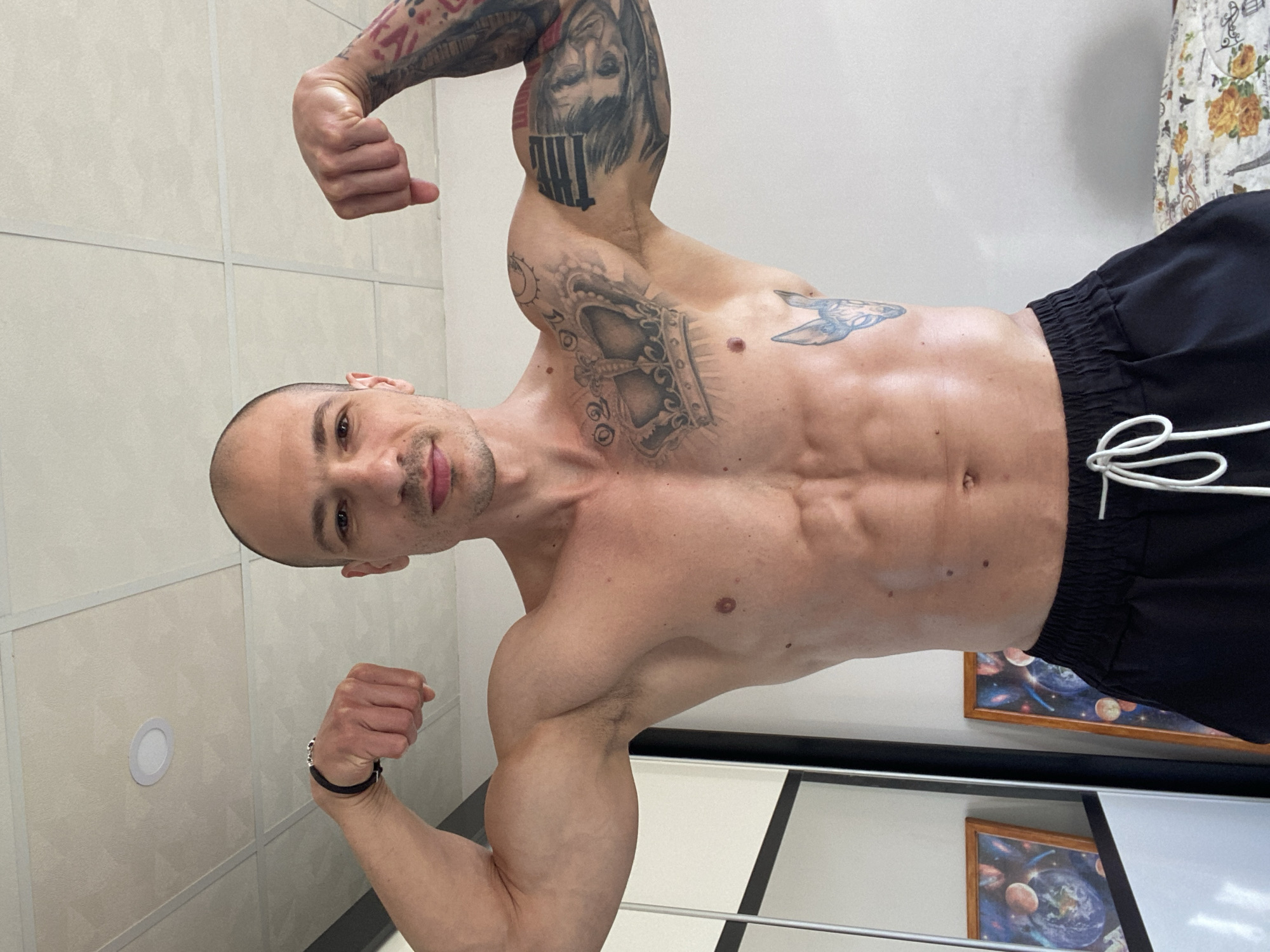 A muscular, bald man is posing indoors, flexing his arms. He is wearing black shorts and has multiple tattoos on his arms, chest, and stomach. The background includes a tiled wall, a painting of space, and a wardrobe with a mirror. The location is not identifiable as the city of Pleven, Bulgaria, from the image alone.