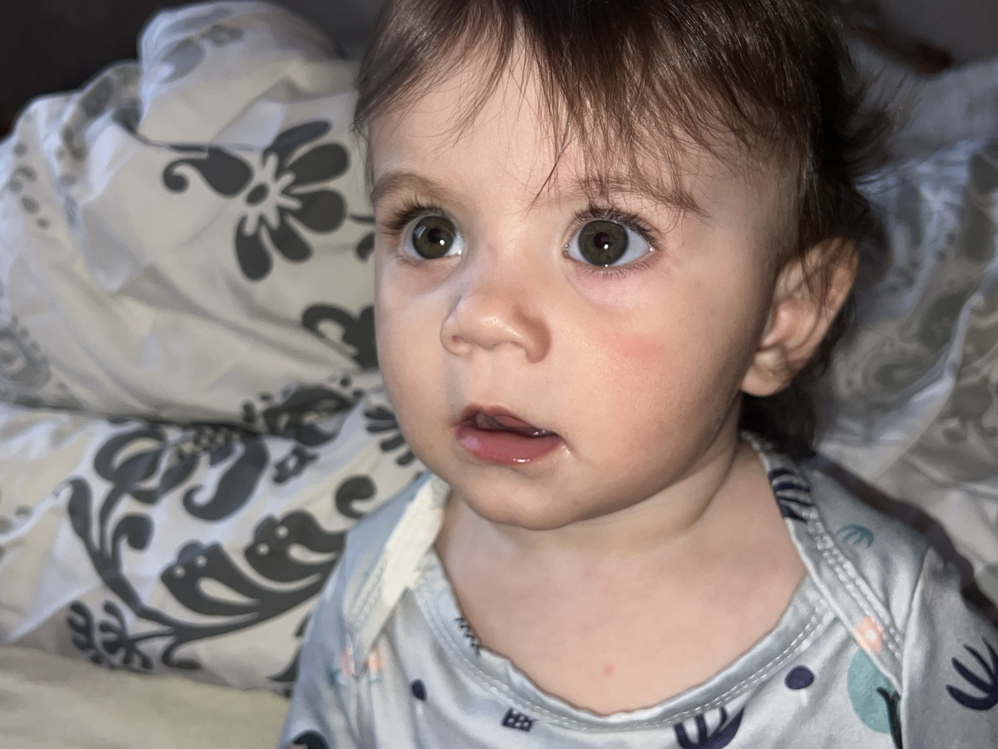 A close-up shot depicts a baby with brown hair and eyes, wearing a light blue and white patterned onesie. The baby is lying on bedding with a gray and white floral pattern. The baby appears to be looking off to the left with a slightly open mouth. The location in the city of Elena, Bulgaria is not discernible from the image.