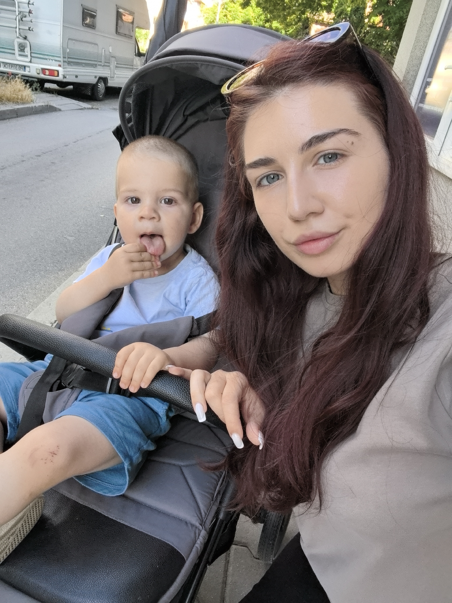 In Stara Zagora, Bulgaria, a woman with long, dark red hair, wearing sunglasses on her head and a beige t-shirt, takes a selfie outdoors. She is seated next to a baby in a stroller. The baby, wearing a light blue t-shirt and blue shorts, is facing the camera with its tongue sticking out and its hand near its mouth. The stroller has a black handle and grey padding. In the background, a motorhome is parked on a street next to a sidewalk. Trees and a building are also visible. The lighting suggests it is daytime.