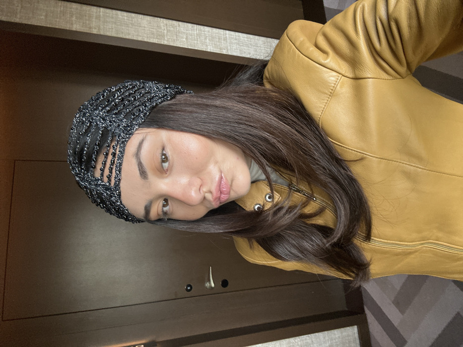 A female subject, appearing to be in an indoor setting, captures a head-and-torso selfie. She has dark brown hair, brown eyes, and is depicted with pouting lips. She wears a black, textured or beaded headpiece resembling a turban, and a mustard yellow leather jacket with a zippered front closure and silver-toned snaps on the collar. A light-colored turtleneck or high-collar garment is visible beneath the jacket. The background features a dark brown, plain wooden door with a silver-colored handle and a black circular lock on the left, suggesting a hotel room or private residence. To the right, vertical paneling with a subtle texture is visible. The floor beneath the subject is partially visible, showing a geometric patterned carpet in shades of gray and brown. The scene is understood to be within the city of Porto, Portugal, likely in a modern residential or hospitality interior.