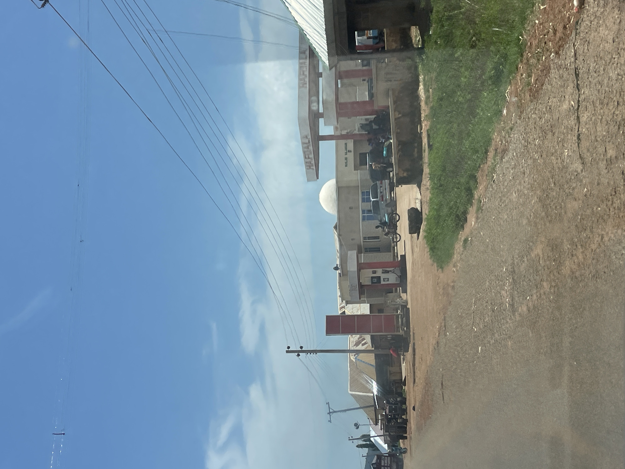 The image shows a street scene in Zalau, Nigeria, with a prominent gas station. The gas station has a red and white structure with a canopy and signage that reads "HAFDALA". Several vehicles are present, including a motorcycle, a car, and what appear to be tractors. People are seen standing and interacting near the gas station. The surrounding area features a mix of buildings with corrugated metal roofs, suggesting a developing urban environment. Overhead power lines crisscross the sky, which is blue with scattered clouds. The road appears to be unpaved or partially paved, with a dirt shoulder and some vegetation. The overall impression is of a functional, bustling street in a Nigerian town.