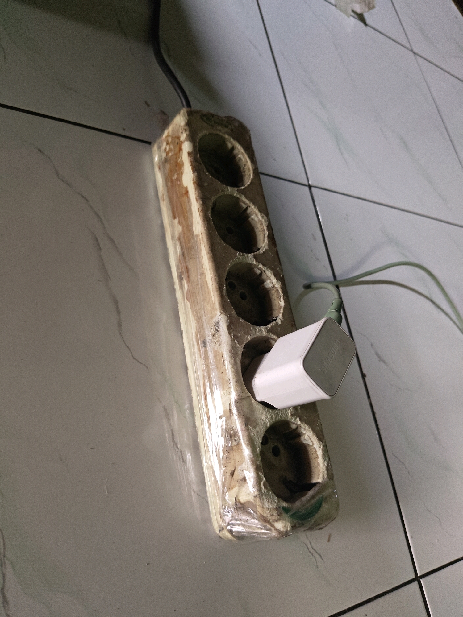 In a tiled room in Sumberwono, Indonesia, a close-up shot captures a power strip with multiple sockets, showing signs of damage and wear. One socket is occupied by a white Samsung power adapter plugged into a green cable. The power strip itself appears to be constructed from a wooden block, wrapped in clear plastic, with visible degradation and openings for the electrical outlets. The tiled floor, a common feature in Indonesian homes, is visible in the background, with grout lines creating a grid pattern. The lighting suggests an indoor environment, possibly a home or an office. The image focuses on the power strip and its immediate surroundings, hinting at a functional, though not pristine, electrical setup.