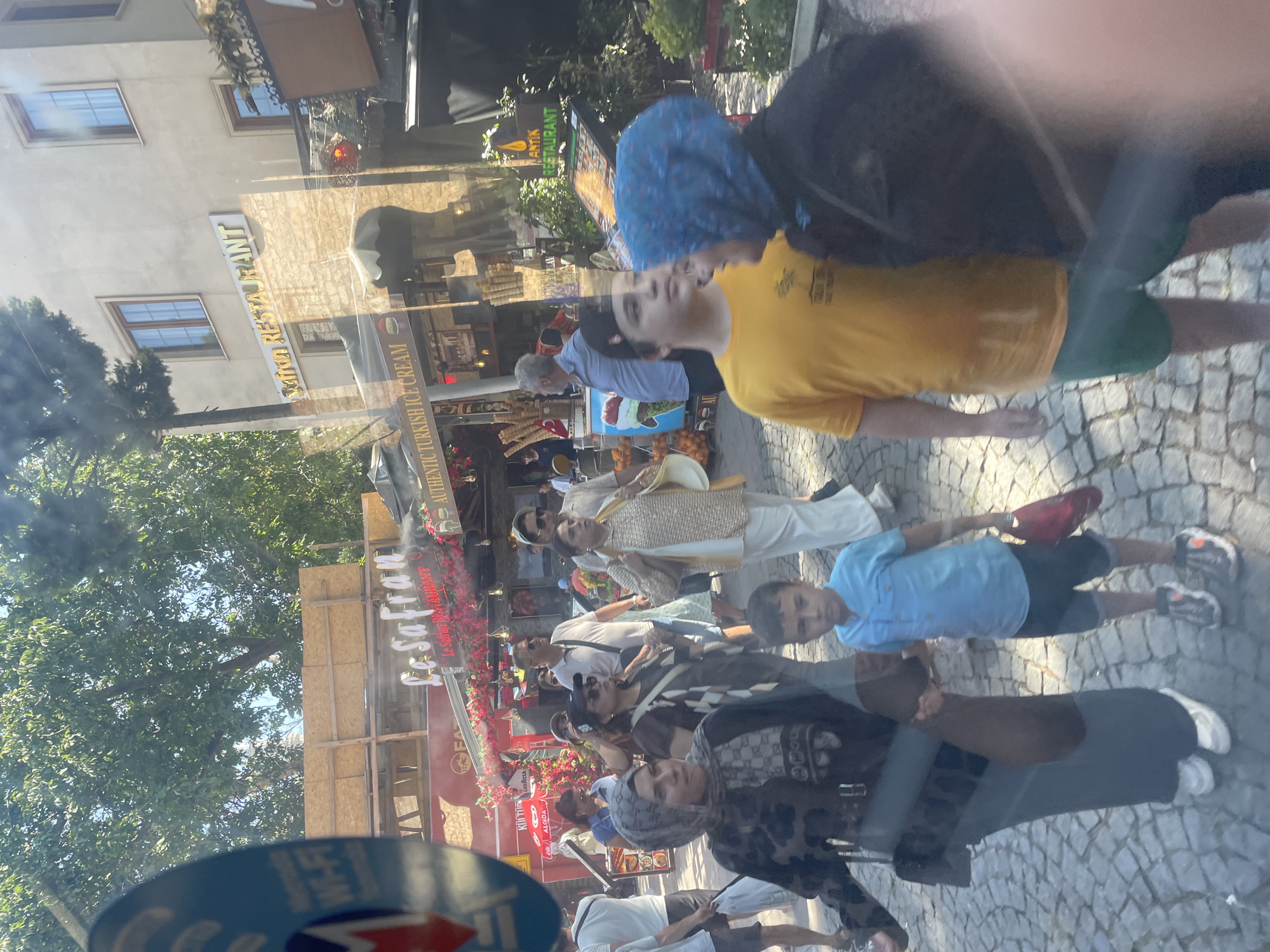 In Istanbul, Turkey, a bustling street scene unfolds under a clear sky, with lush green trees providing dappled shade over a cobblestone path. Several individuals are visible, some walking, others observing. A young boy in a bright blue shirt and dark shorts walks with a woman in a black patterned dress and headscarf, his right hand holding hers. Nearby, a young man in a yellow t-shirt and green shorts stands, his gaze directed elsewhere. Behind them, a woman with a blue head covering and dark jacket is partially visible. Storefronts line the street, featuring signs like "Le Safran Restaurant" and "Authentic Turkish Ice Cream," adorned with colorful decorations. Oranges are stacked near an ice cream stand, suggesting a vibrant marketplace atmosphere. The overall impression is one of a lively, sun-drenched day in a historic urban setting.