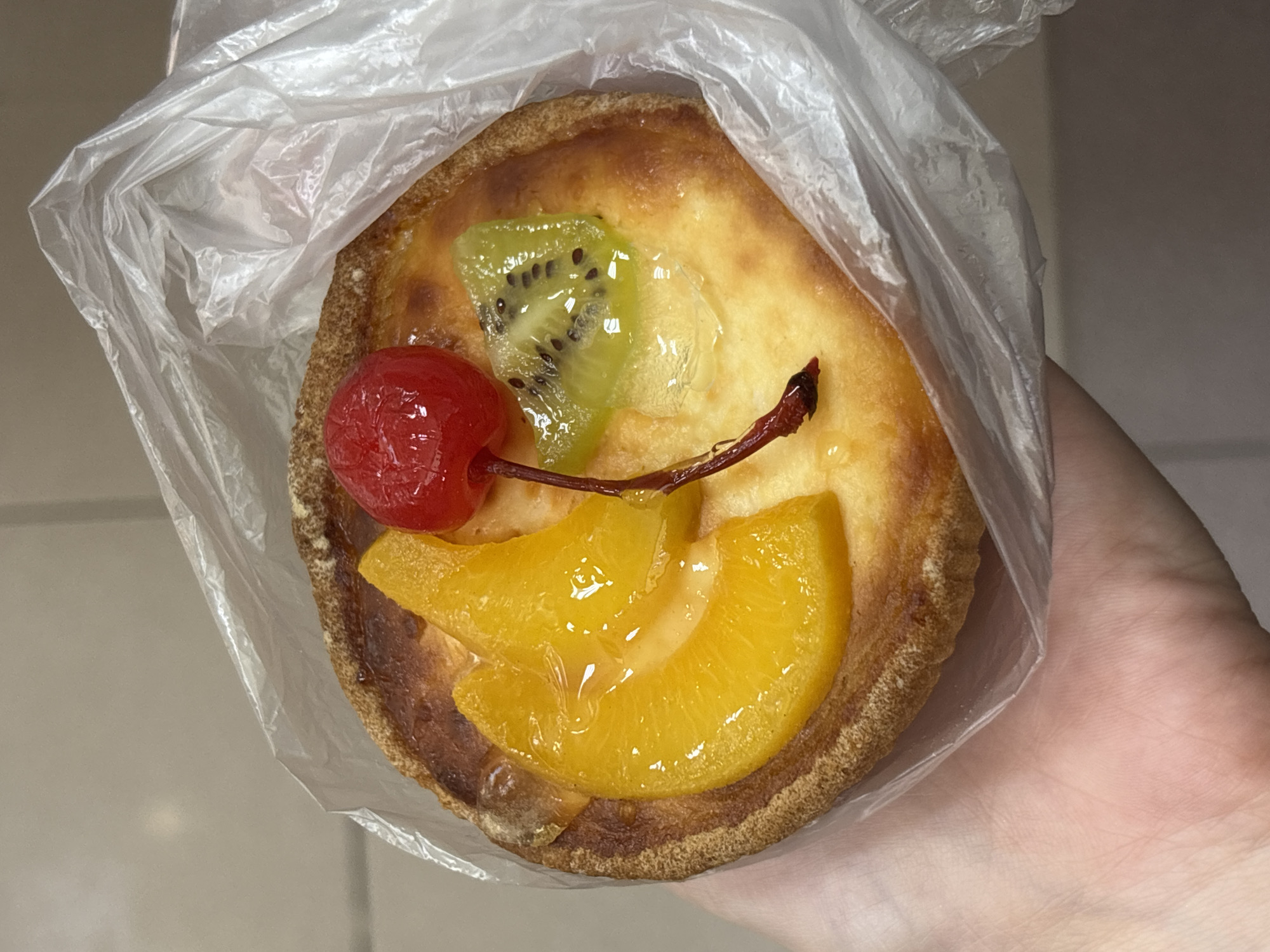 A hand holds a fruit tart in a plastic bag. The tart has a golden brown crust and is topped with sliced peaches, a slice of kiwi, and a maraschino cherry. The background is a plain, light-colored surface, possibly a wall or countertop. The image does not provide any information about the location in the city of Chaikovsky, Russia.