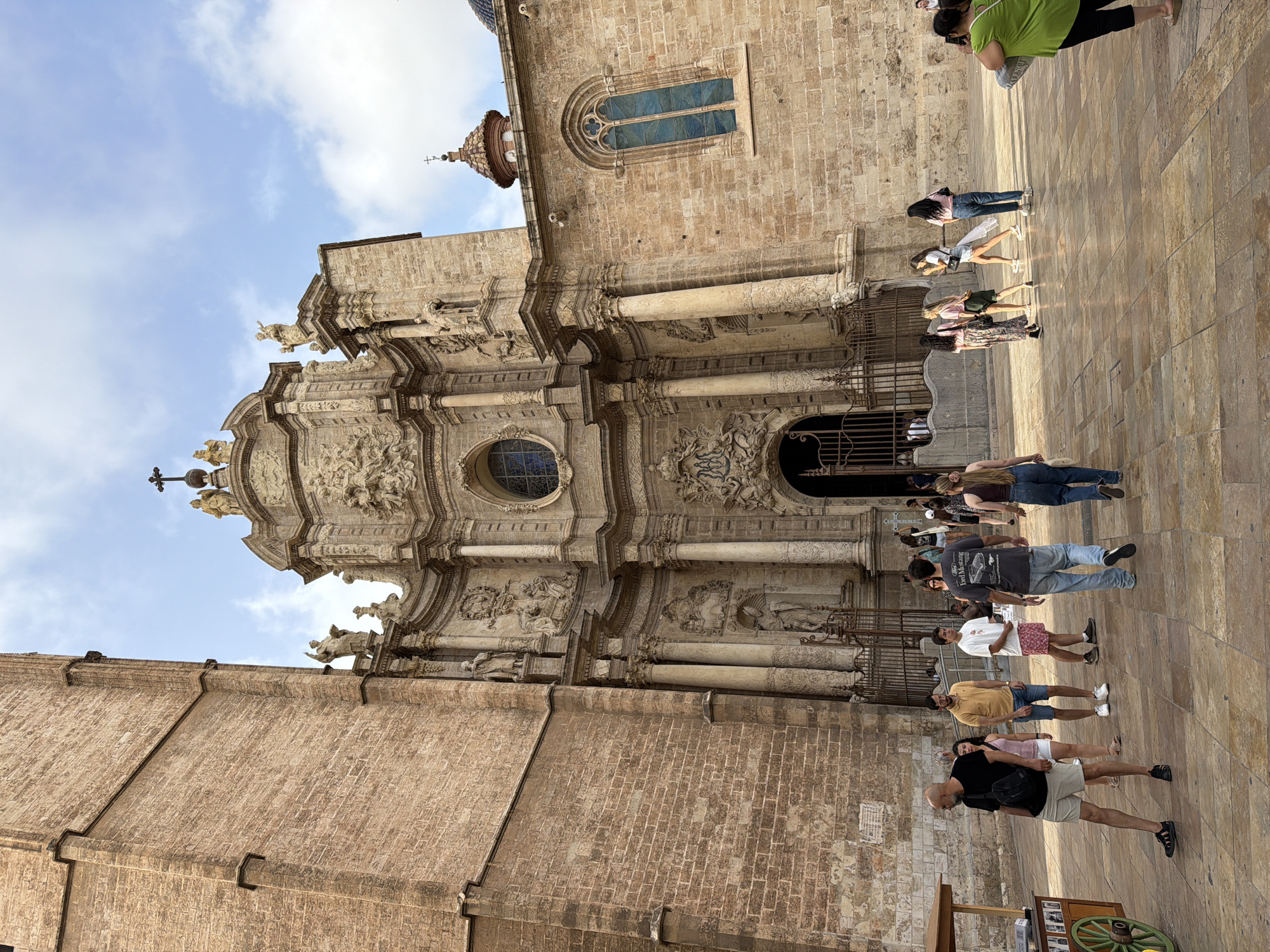 In front of the Valencia Cathedral, several people are walking and standing on a paved plaza. The cathedral is made of weathered stone with intricate carvings and large arched doorways. A tall, brick tower is visible to the left of the cathedral. A street cart with a green wheel is on the far left. The sky is partly cloudy.