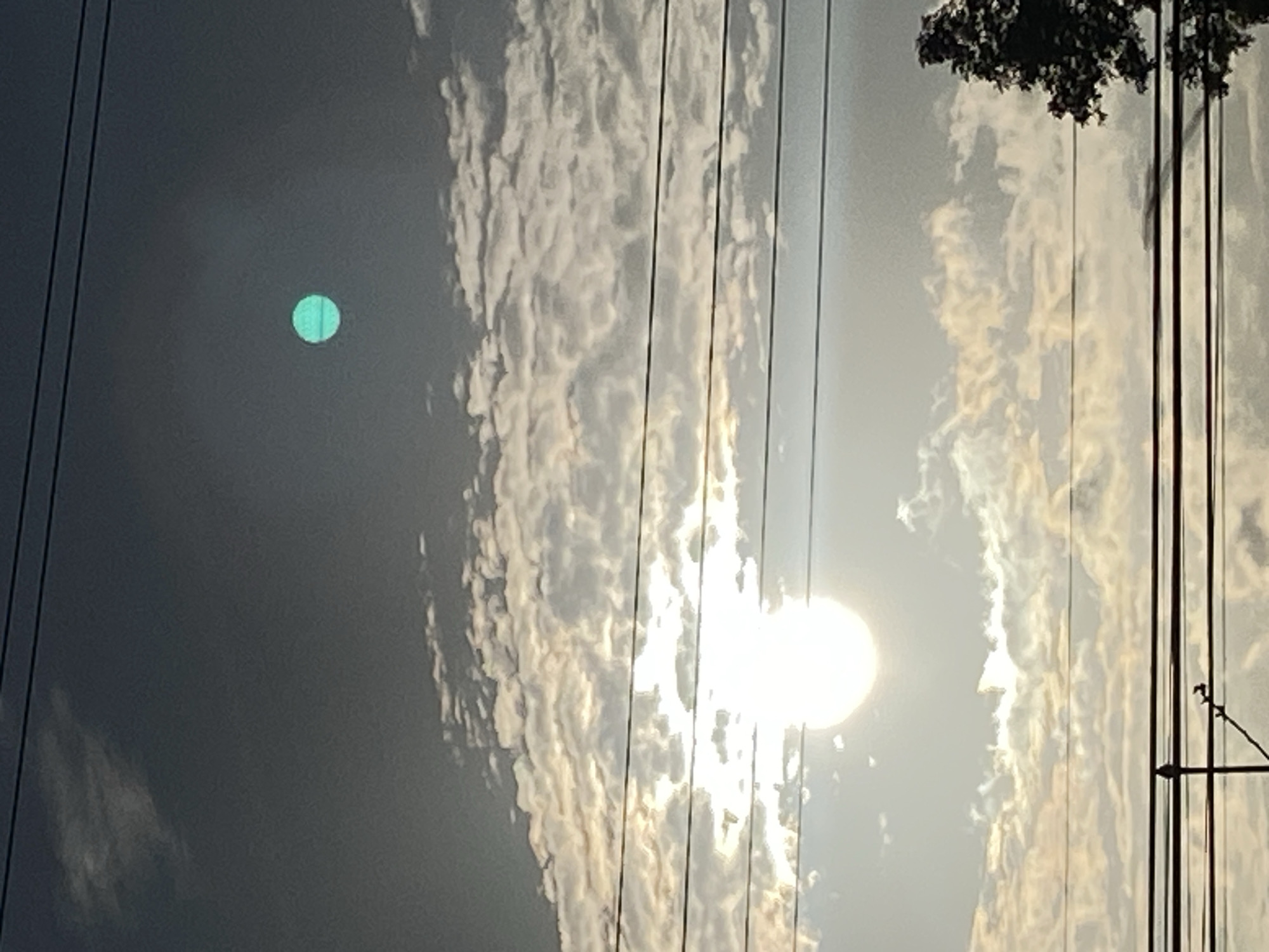 The image shows a daytime sky in Tinaquillo, Venezuela, with a bright sun obscured by clouds. Power lines are visible across the frame, and a tree is in the upper right corner. A green spherical object is in the upper left.