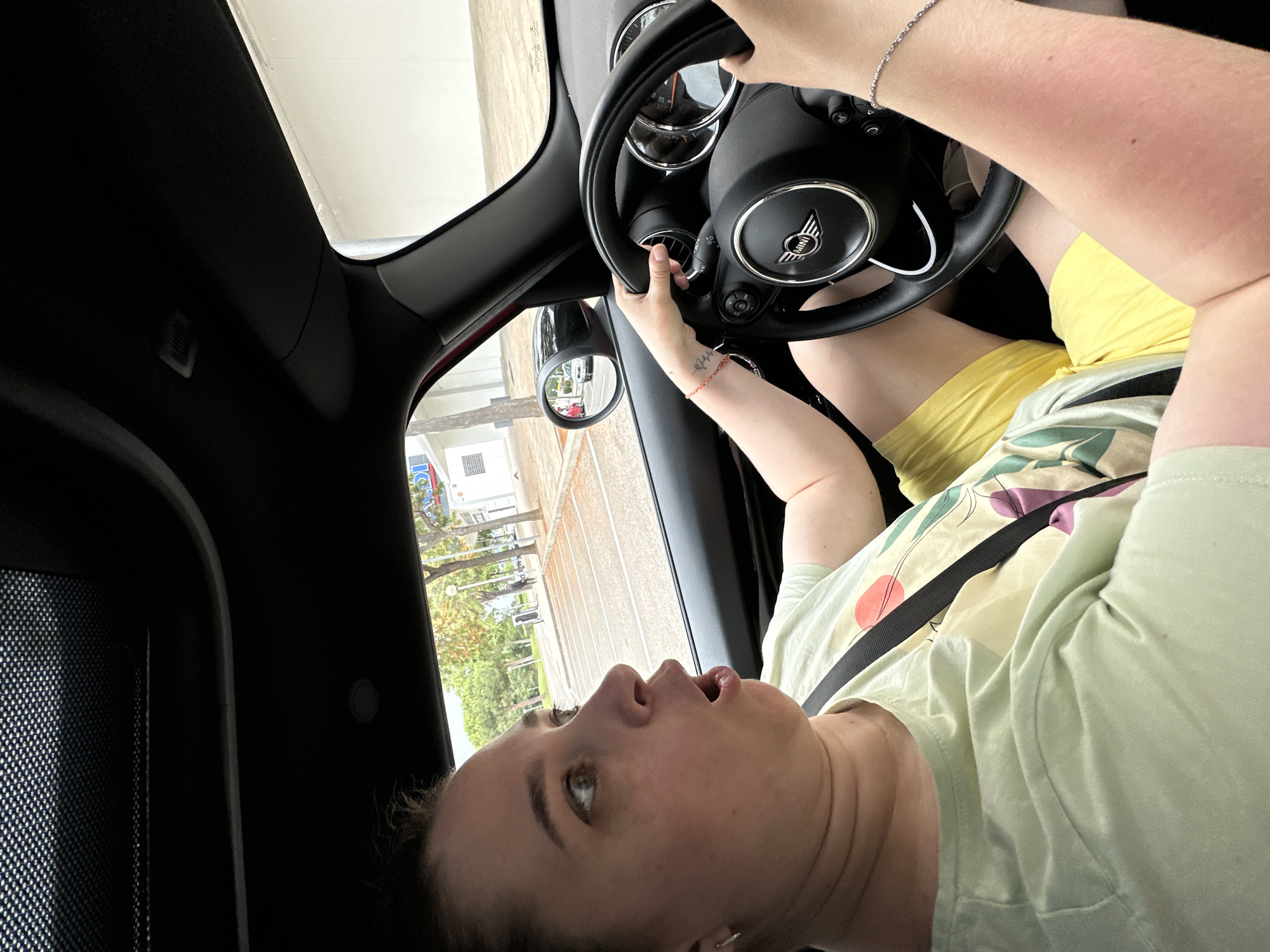 A woman is seated in the driver's seat of a Mini Cooper, holding the steering wheel with both hands. Her mouth is open in a surprised expression, and her eyes are wide, looking to the left. She is wearing a light green t-shirt with a colorful abstract print and yellow shorts. A black seatbelt is visible across her chest. The car's interior is dark, with a portion of the sunroof visible at the top of the frame. Outside the car, a parking lot with painted lines is visible, along with trees and buildings. The scene appears to be in Castell d'Aro, Platja d'Aro i s'Agaró, Spain.