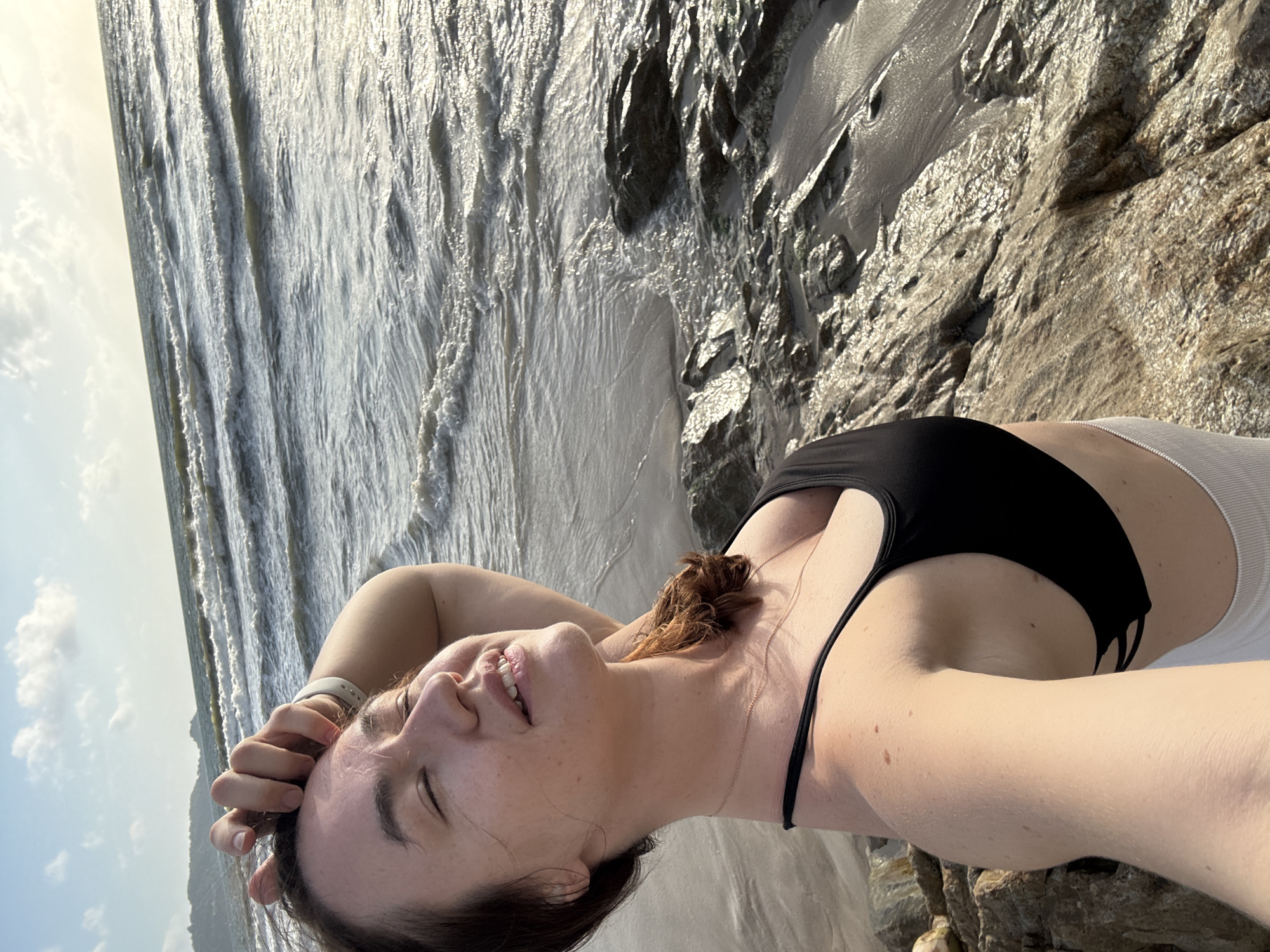 A woman is taking a selfie on a rocky beach in Patong, Thailand. She has her hand on her forehead and her eyes are closed as she smiles. She is wearing a black bikini top and white shorts. The ocean is behind her with waves crashing on the shore. The sky is cloudy.