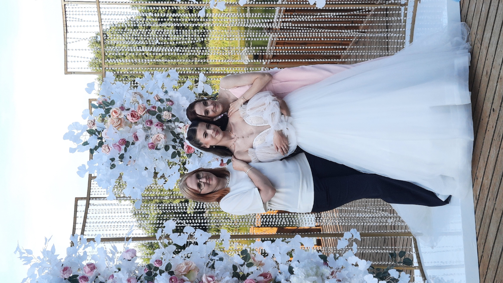 Three women are posing for a photograph in front of a wedding arch decorated with white flowers and crystal strands. The woman in the center is wearing a white wedding dress with a plunging neckline and puffy sleeves. The woman to her left is wearing a white t-shirt and black pants, and the woman to her right is wearing a pink slip dress. The wedding arch is set up outdoors, with trees and a body of water visible in the background. The ground appears to be made of wooden planks. The location is the village of Zhodzishkavets, within the Zhodzishkavetsky Selsoviet, Belarus.