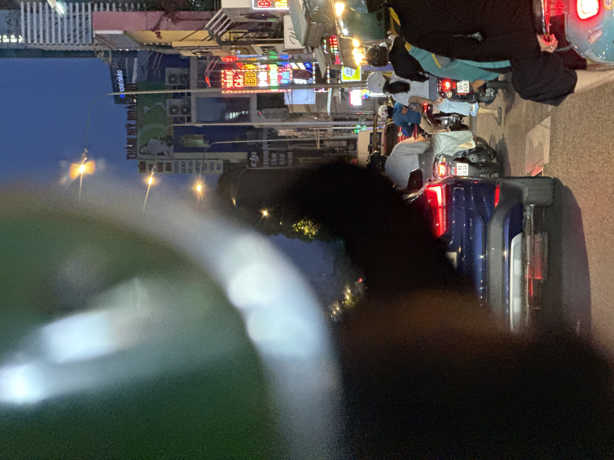 The image depicts a street scene at night in Thủ Đức, Vietnam. A row of vehicles, including cars and motorbikes, is shown in a line of traffic, oriented vertically in the frame. Streetlights illuminate the area, and neon signs adorn the buildings lining the street, displaying a variety of advertisements and names of businesses. The signs include the name "THÁNH MỸ HOÀ," advertising "hotel nikko", the name "ED" and "PET SHO" and "LITTLE." The license plates are visible, the first one is "9-NA 08.06" and the other is "59-A 848.9". A blurred object is in the foreground and obstructs part of the view.