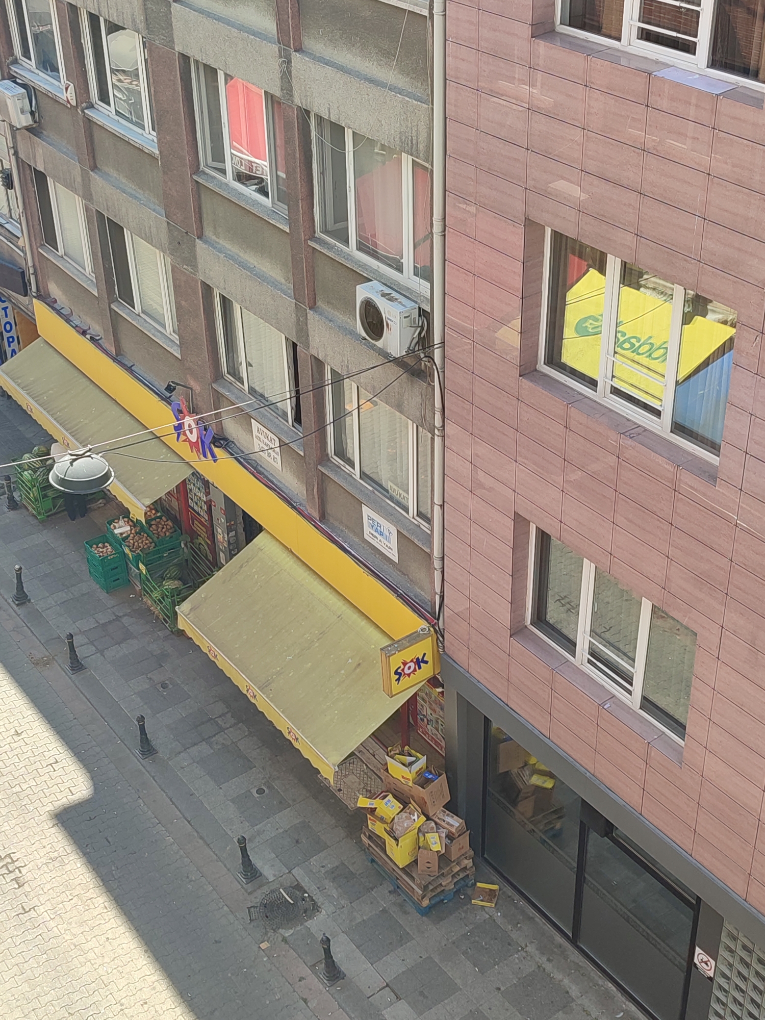The image depicts a street scene in Kadıköy, Turkey, viewed from an elevated perspective. In the foreground, a tiled pedestrian walkway runs alongside a street. Several black bollards are spaced along the edge of the walkway. To the right, a building with a pink tiled facade features large windows, one of which reflects a bright yellow surface with green lettering that appears to be a logo. Below this, another window reflects the interior of the building.
Adjacent to the pink-tiled building is a gray-colored building with multiple windows and air conditioning units mounted on its facade. A yellow awning extends from the front of this building, covering the entrance to a shop. The shop appears to be a "ŞOK" market, indicated by signage. Outside the shop, green crates are filled with produce, including watermelons and potatoes. Stacks of cardboard boxes are piled on wooden pallets near the entrance. A streetlamp hangs above the awning. The overall scene suggests a commercial area with a market operating on a street level.