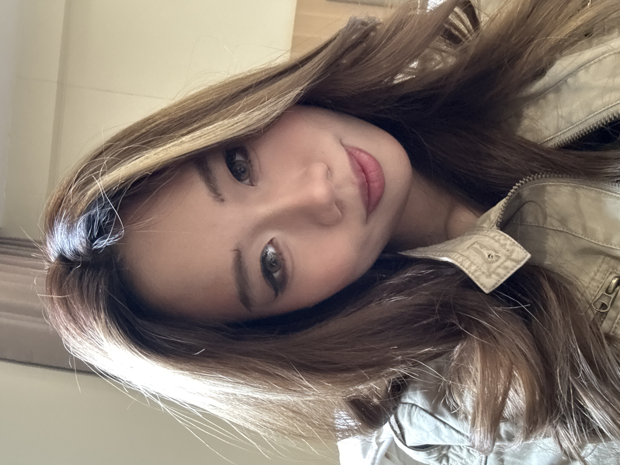 The image is a close-up, slightly angled selfie of a woman with long, light brown hair. Her hair has blonde highlights and frames her face. She has light skin, brown eyes, and pink lips. She appears to be wearing a light-colored jacket. The background is blurry and indistinct. There is no evidence in the image to suggest the location is Dunhuang City, China.