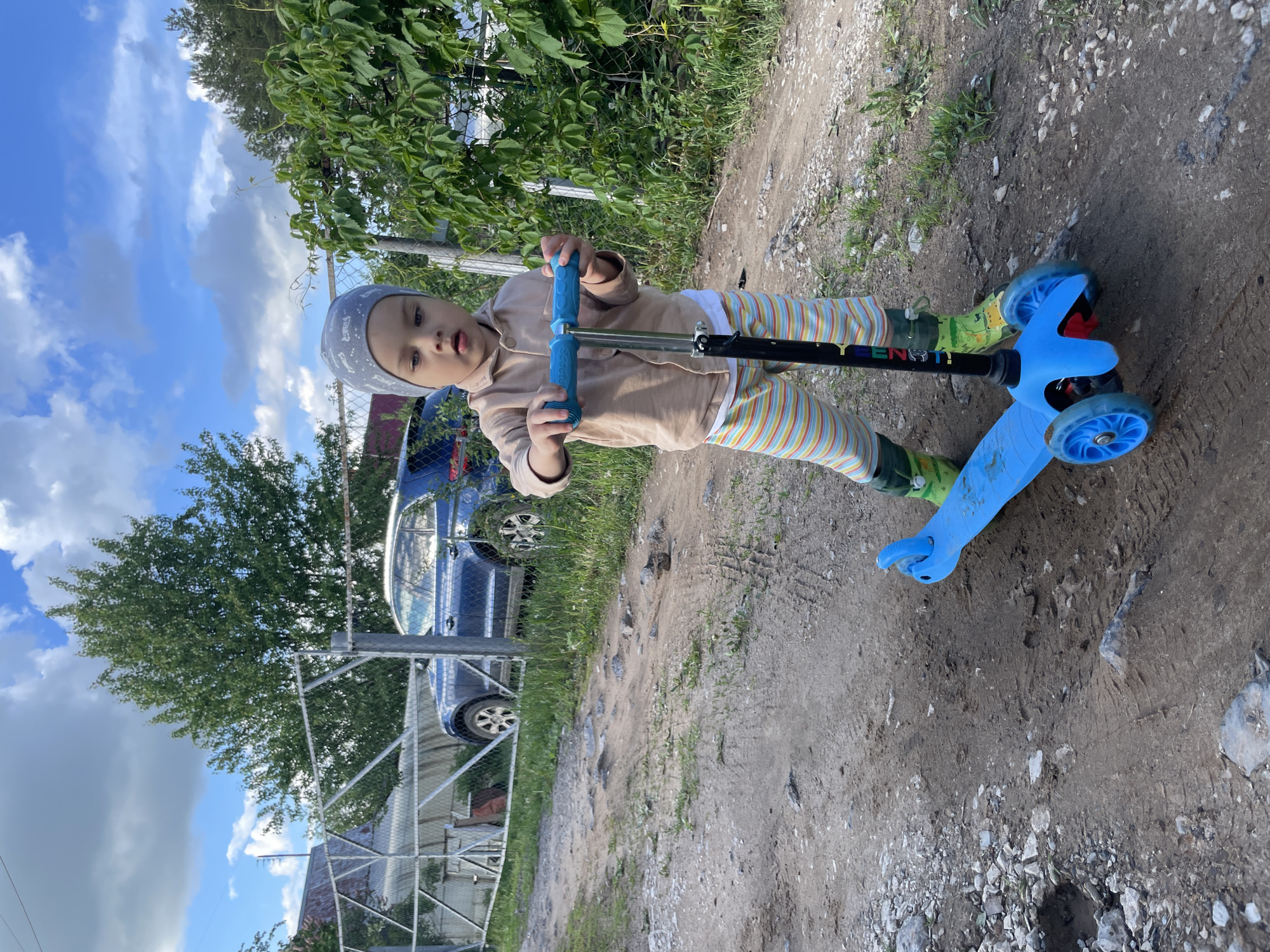 A child wearing a grey hat, beige top, and striped pants stands on a blue scooter on a dirt path. The path has patches of grass and rocks. A chain-link fence runs along the left side of the image, with a blue car visible behind it. Green foliage is present above the fence, and a blue sky with white clouds is visible. The location is likely Zav'yalovo, Russia.