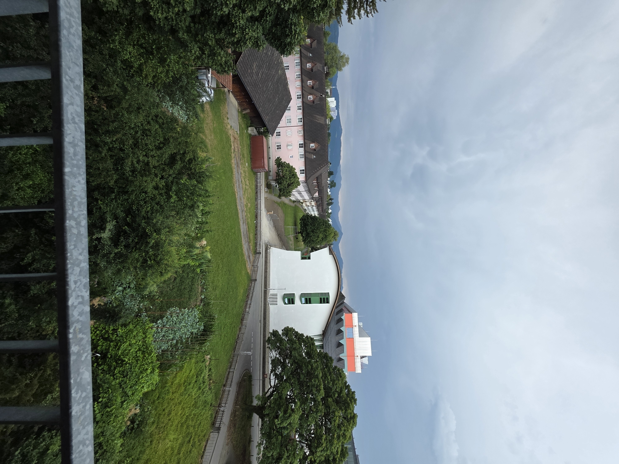 This is a vertically oriented photograph taken from an elevated vantage point, looking out over a landscape that appears to be in Bregenz, Austria. The foreground is dominated by a grey metal railing, suggesting the viewer is on a balcony, bridge, or elevated walkway.
The scene depicts a blend of urban and natural elements. To the left, lush green trees and vegetation line a grassy slope. Further back, a long, multi-story pink building with a dark roof and many windows is visible, interspersed with other structures, including a barn-like building with a wooden exterior and a dark tiled roof. A shipping container is also situated near this building.
In the mid-ground, a modern white building with a distinctive curved roof and bright green window frames stands out. Adjacent to it is another building with a grey roof and colorful orange and red architectural elements. A large, mature tree with dense foliage is prominent in front of this modern structure. A road runs alongside the buildings, with a few indistinct figures potentially visible on it or on pathways.
The background features rolling hills or mountains shrouded in a hazy atmosphere, characteristic of a somewhat overcast day. The sky above is a mix of grey and light blue clouds, indicating a cloudy or potentially rainy day. The overall impression is one of a semi-urbanized area nestled within a natural environment. No specific activity or event is clearly discernible, and no text is visible in the image. The time of day appears to be daytime, given the natural light.