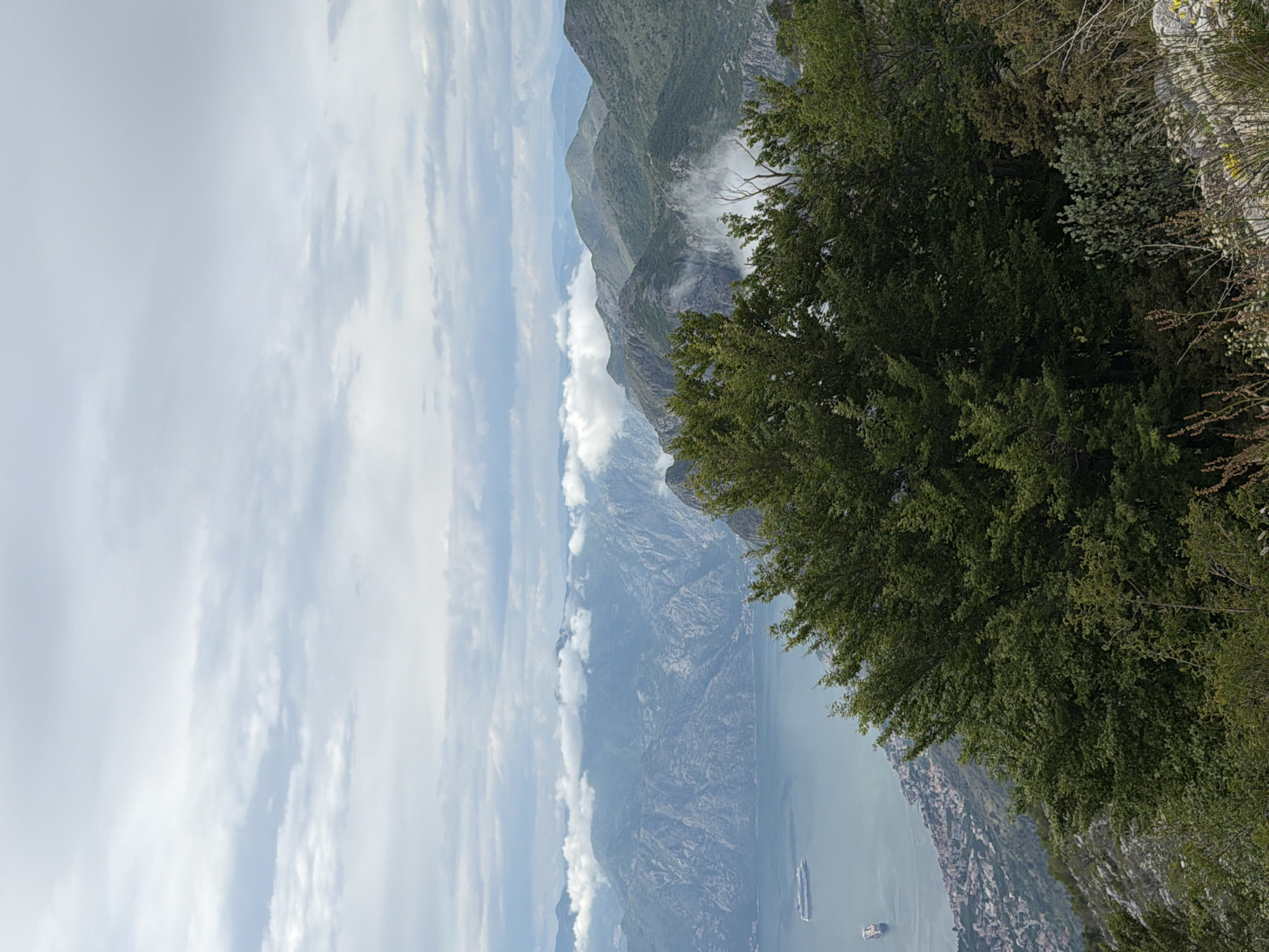 The image depicts a mountainous landscape from a high vantage point, likely near Žanjev Do, Montenegro. A body of water stretches into the distance, with two vessels visible. Clouds are present in the sky and also envelop portions of the mountain range. The foreground features dense green foliage.