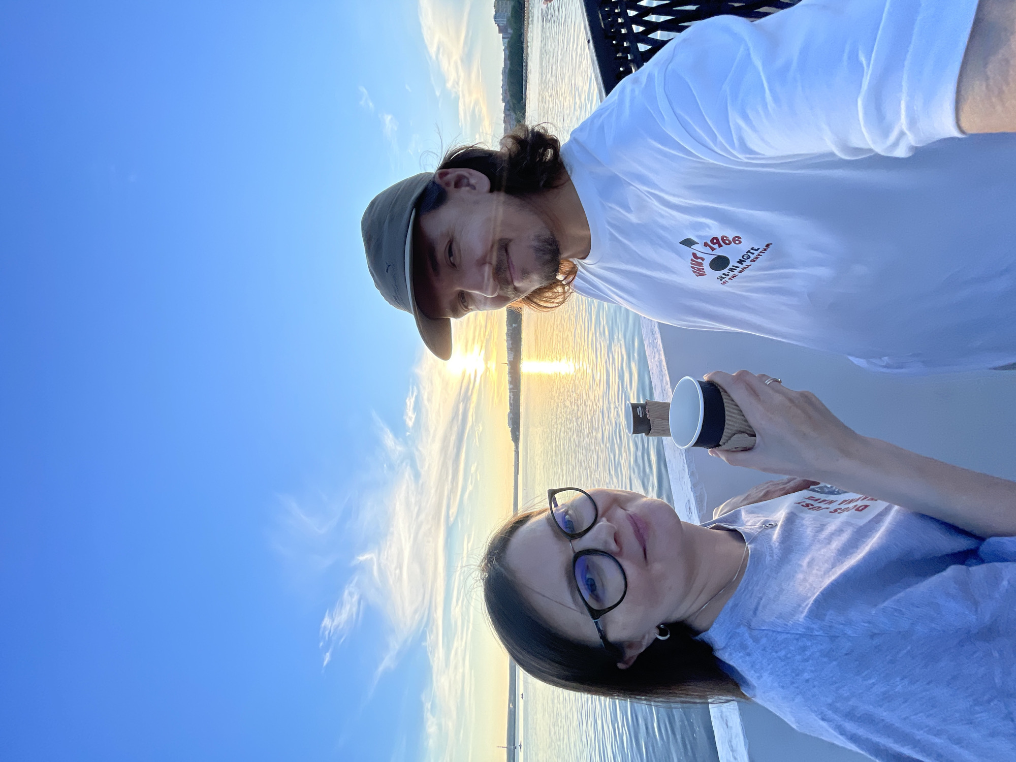 An outdoor, eye-level, full-body shot captures a smiling couple standing on a walkway with a body of water and a sunset behind them. The woman on the left wears glasses, a light blue t-shirt, and hoop earrings. She holds a paper coffee cup in her right hand. The man on the right wears a light green cap and a white t-shirt with a red and black graphic on the front. He has a mustache and goatee and long, dark hair. The water reflects the sunset, which is partially obscured by clouds. In the distance, buildings line the shore.