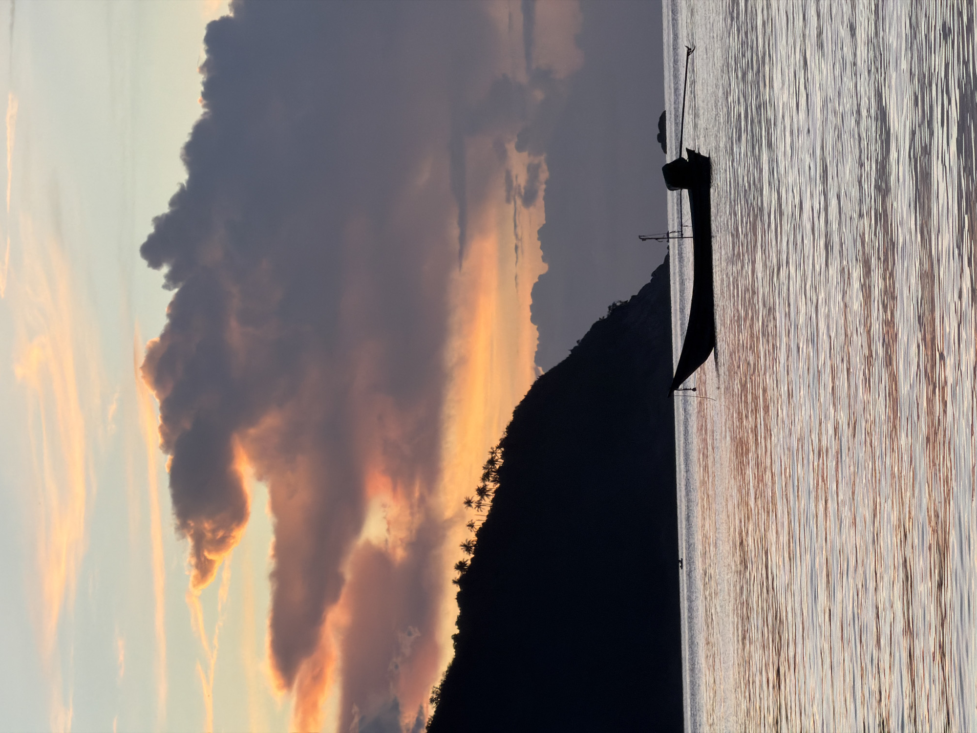 A silhouette of a long-tail boat floats on calm, rippling water. The boat is dark, with an outboard motor visible at the stern and a rope tethered to its bow. In the background, a dark, tree-covered island rises from the sea, with a line of palm trees silhouetted along its ridge. Above the island, the sky is a dramatic canvas of sunset colors, with vibrant orange and pink hues mixing with deep purples and grays in the clouds. The water reflects these colors, creating a soft, shimmering effect. The scene evokes a peaceful and serene atmosphere, characteristic of a sunset on Koh Phangan island, Thailand.