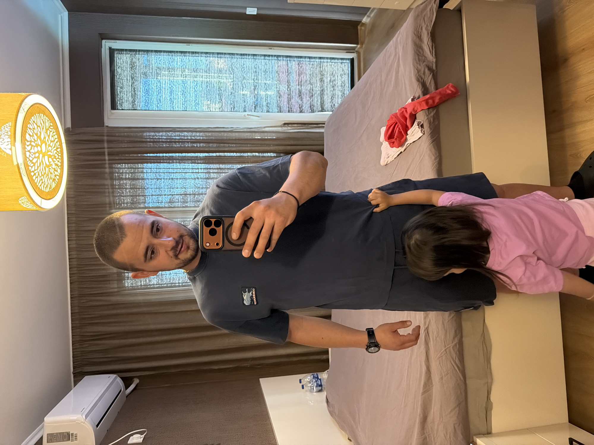 In this media file, a man and a young child are captured in a domestic setting, likely a bedroom. The man is taking a selfie with his phone, holding it in his right hand. He is wearing a dark blue t-shirt and dark shorts, and a black watch on his left wrist. The child, wearing a pink t-shirt, lies on a bed next to him, facing away from the camera. A pile of red and white clothing rests on the bed between them.
The room features a window with blinds, letting in natural light, suggesting it could be daytime. A lamp with a decorative, tree-like design is visible on the left side of the frame. An air conditioning unit is mounted on the wall near the floor in the lower left corner of the image. The flooring appears to be wood. The overall mood is casual and intimate, hinting at a moment of everyday life. No specific time of day or weather conditions are discernible beyond the natural light. There is no visible text in the image. The context provided places the location as Burgas, Bulgaria.