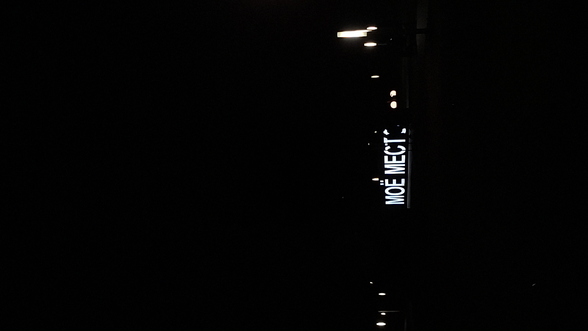 A lighted sign displaying "MÖE MECT" in white letters against a black background is visible in the lower center of the image. The sign is illuminated, making the text stand out in the darkness. Surrounding the sign, there are faint lights in the background, suggesting an urban environment at night. The scene is set in Myadzyel, Belarus.