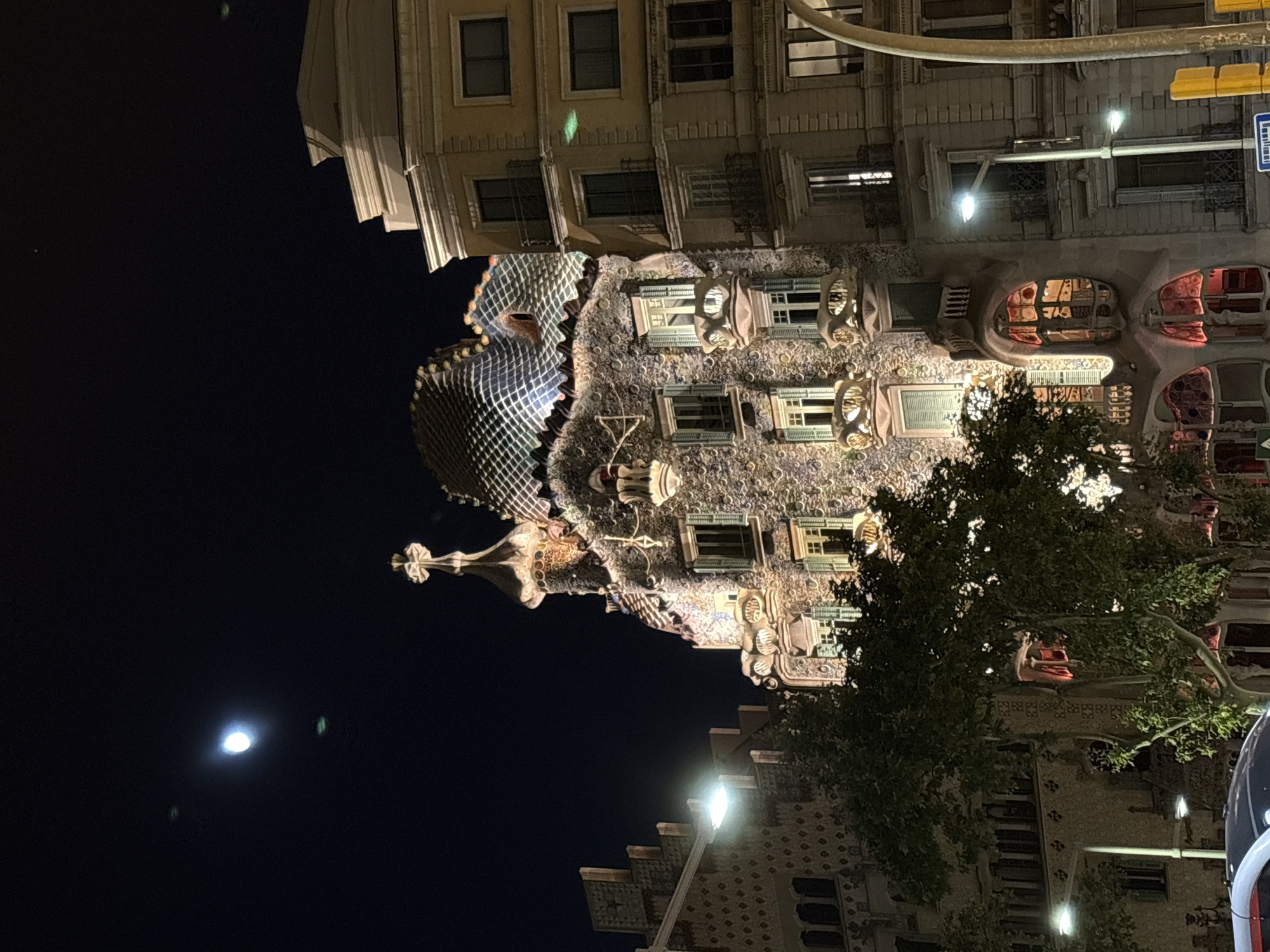 The image shows a nighttime view of Casa Batlló, a modernist building designed by Antoni Gaudí, located on Passeig de Gràcia in Barcelona, Spain. The building's facade is illuminated, showcasing its unique organic forms and colorful mosaic tiles. The roof is particularly striking with its scaled appearance, resembling a dragon's back, and crowned with a cross.
To the right of Casa Batlló, another building with a more traditional architectural style stands, its facade also lit up, revealing rows of windows and balconies. A large tree with dark green foliage partially obscures the lower section of Casa Batlló on the left side of the image. The sky above is dark, with a bright full moon visible in the upper left quadrant, casting a soft glow. Streetlights are also present, illuminating parts of the street and sidewalks. A traffic light with yellow and blue signals is visible on the right side of the frame. The scene appears to be a street or pedestrian area, with elements suggesting urban infrastructure. No people are clearly visible in the image.