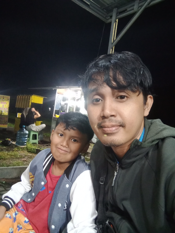 This nighttime image, set in Turunsih, Indonesia, centers on two individuals in the foreground. On the right, an adult male with dark, wavy hair, a mustache, and light stubble wears a dark green hooded jacket. A faint yellowish residue is visible on his lower lip. He directly faces the camera, holding it, indicating a selfie. To his left, a young male child, approximately 8-12 years old, with short dark hair, smiles slightly. He wears a red t-shirt beneath a blue and white letterman-style jacket and patterned orange pants. In the background, under dim artificial lighting, a street vendor setup is visible. This includes a yellow corrugated metal kiosk on the left, adjacent to a white counter. A yellow sign with partial Indonesian text, including 'CIDE CIDE', is affixed to the left of the kiosk. Two additional, indistinct figures are present in the mid-ground, one seated on a light green plastic stool beside a large blue water cooler bottle. An overhead metal structure is partially visible in the upper frame. The immediate ground appears to be a mix of dirt and sparse vegetation.