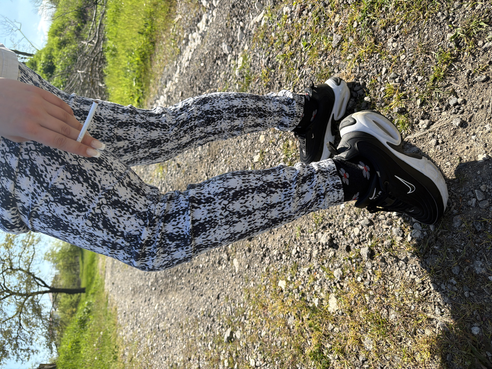 A person wearing black-and-white patterned leggings and black Nike sneakers is sitting on a gravel path. They are holding a lit cigarette in their hand. The surrounding environment consists of a gravel path and sparse greenery. The image was taken in Priseltsi, Bulgaria.