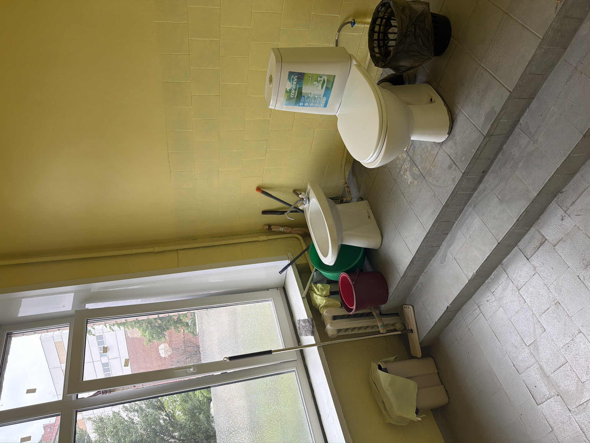 The image depicts a bathroom area with two toilets and a bidet. The room has yellow tiled walls and a large window. Cleaning supplies, including mops, buckets, and rags, are visible. A trash can is located next to one of the toilets. The overall impression is of a functional, possibly older, public restroom. The image does not contain any information indicating its specific location in Kyiv, Ukraine, or the presence of any people or actions.