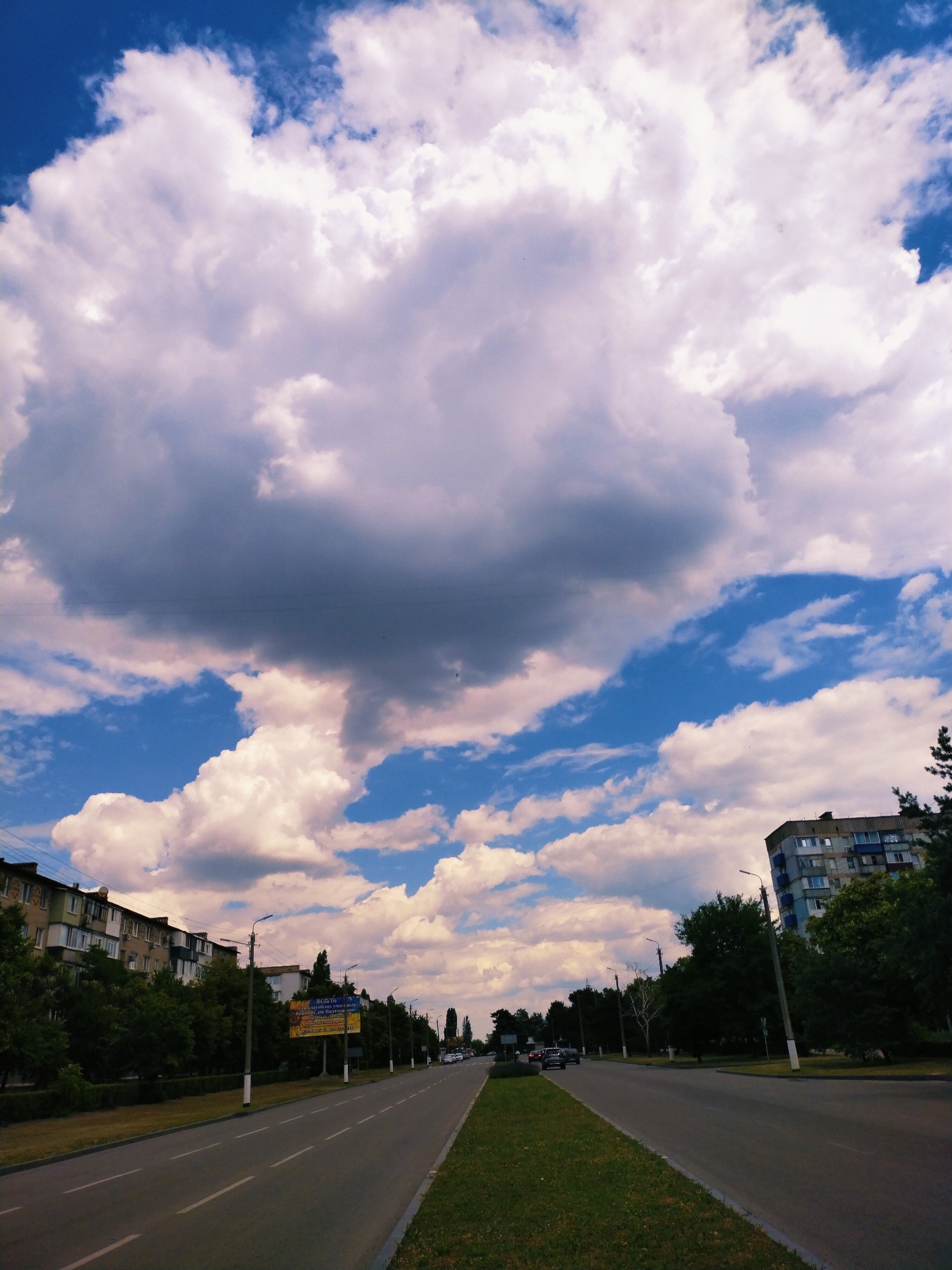 The image depicts a wide, paved road in Horishni Plavni, Ukraine, under a dramatic sky filled with large, fluffy white clouds and patches of blue. The road appears to be a multi-lane highway or a major thoroughfare, with painted lane markings visible. On either side of the road are green grassy verges and rows of trees.
Along the left side of the road, there are multi-story apartment buildings with balconies, typical of Soviet-era architecture, interspersed with dense green foliage. On the right side, the trees are also lush and green, with a tall apartment building visible in the distance. Several streetlights line the road, and a few cars are driving on it, suggesting moderate traffic. One car is parked on the side of the road near a large yellow billboard. The billboard has text and images, some of which appear to be in Ukrainian. The overall scene suggests a daytime setting, likely late afternoon or early evening, given the quality of light and the cloud formations. The weather appears to be fair, with the clouds indicating potential for either sunshine or a passing shower. The mood is serene, with the vast sky dominating the composition, creating a sense of openness and the grandeur of nature.