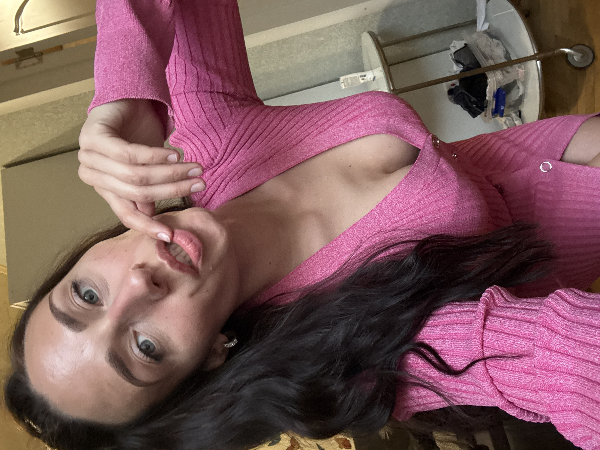 A fair-skinned woman with dark hair and blue eyes is posing in what appears to be an indoor environment. She is wearing a pink ribbed top that reveals a hint of her chest. Her left hand is raised, with her fingers touching her lips. There is an earring visible on her left ear. In the background, there is a metal rack with miscellaneous items. The location is not possible to determine from the image.