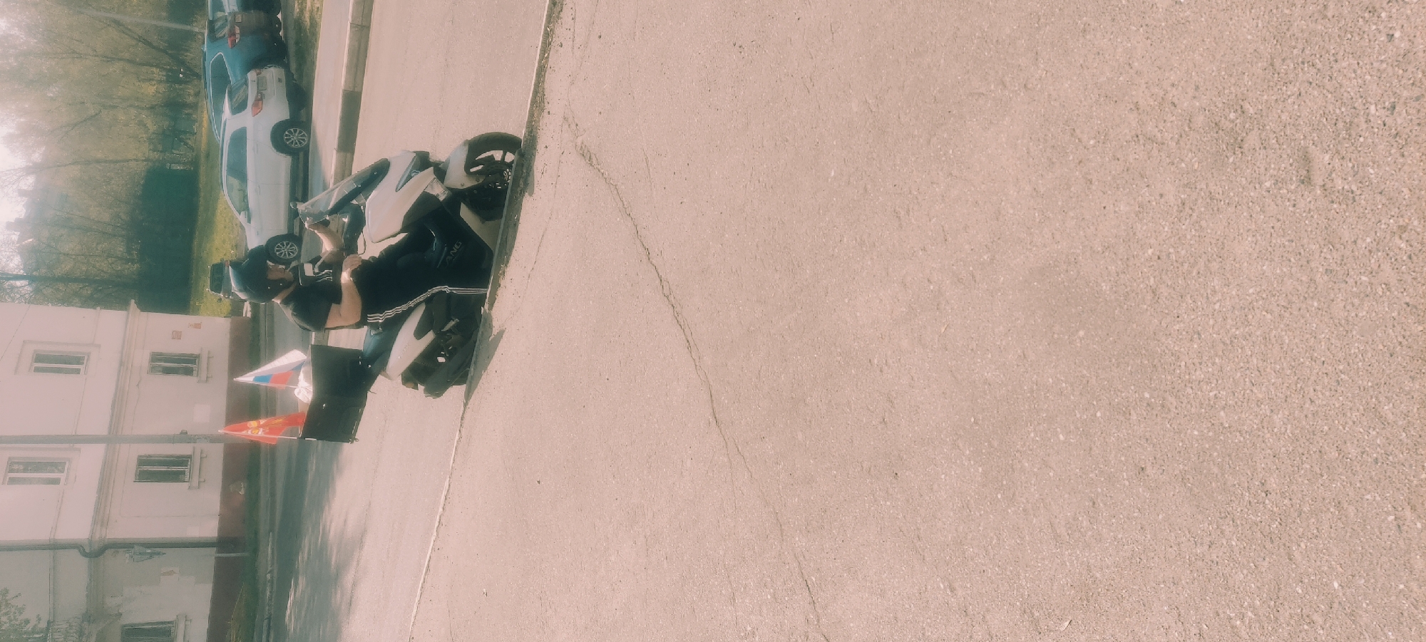 A motorcyclist in Fryazino, Russia is sitting on a white scooter with black accents, facing away from a multistory building. The person is wearing a black t-shirt, black pants with white stripes, a black helmet, and dark shoes. Two small Russian flags are affixed to the scooter, along with a black box. Adjacent to the building, parked vehicles are seen along the side of the road. The scene is set on a sunny day, with the surrounding environment composed of paved streets and some greenery.