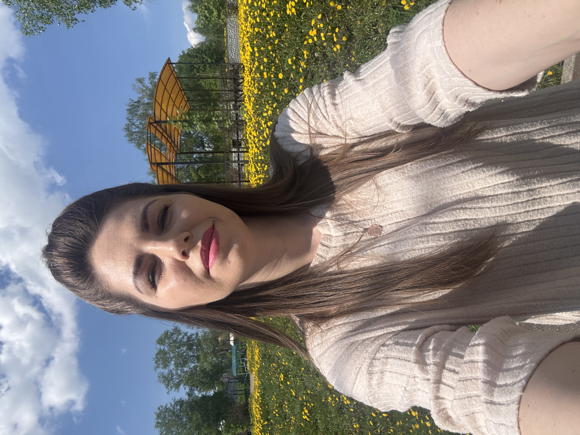 A woman with long brown hair is taking a selfie in a park in Shevlyakovo, Russia. She is wearing a light-colored sweater and pink lipstick. Behind her is a field of yellow flowers and a metal structure. The sky is blue with white clouds.