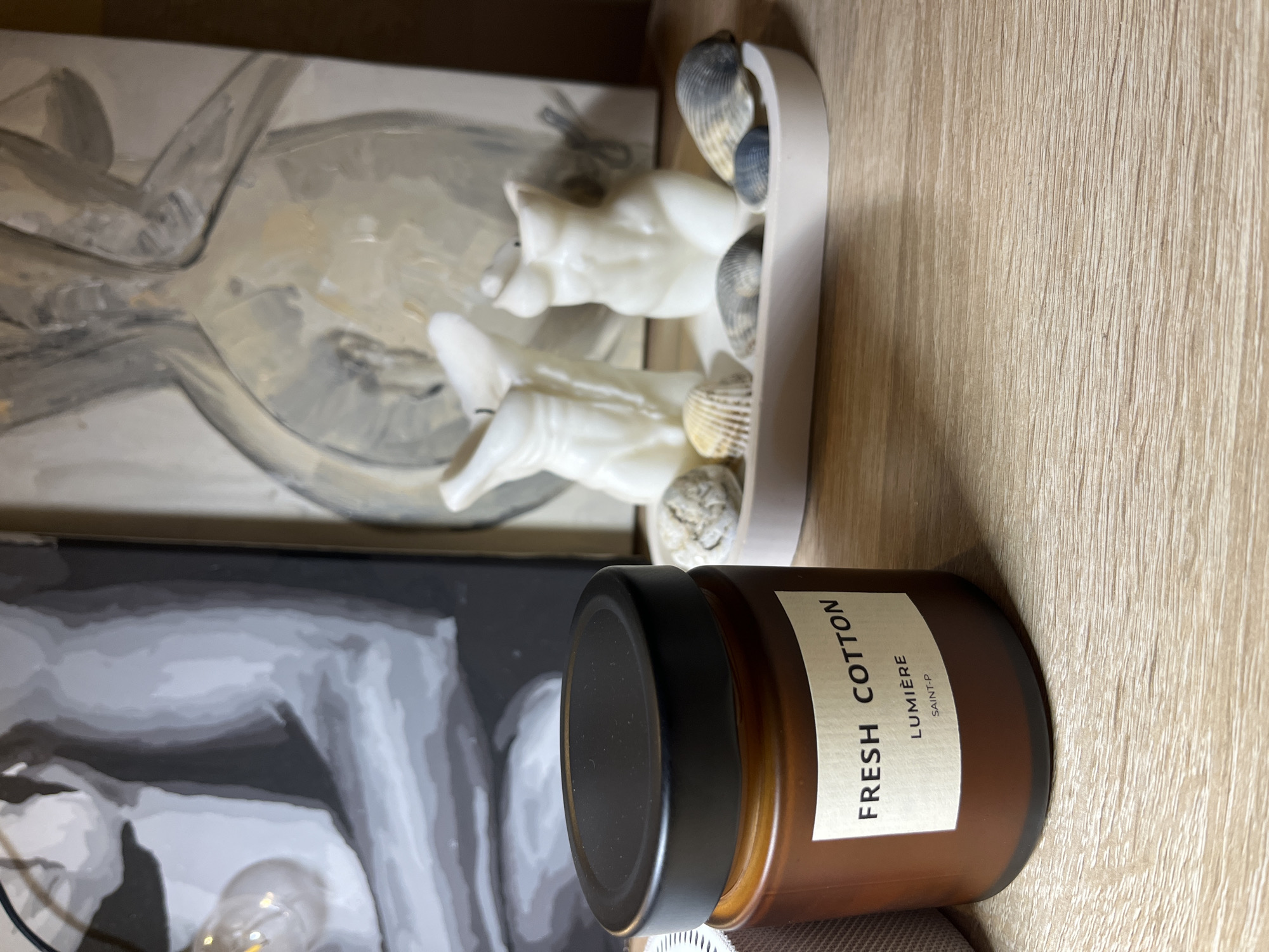 The scene depicts an interior setting with several objects arranged on a light brown wooden surface. An opened jar of "FRESH COTTON LUMIERE SAINT-P" candle with a black lid is in the foreground. Behind it, there's a tray-like object with white figurines and seashells. Adjacent to the tray, there are two paintings with monochrome hues. A partial glimpse of what looks like a light bulb is visible. No people are present. The objects appear to be arranged for decorative purposes. The specific location of Sochi, Russia cannot be confirmed based on the image alone.