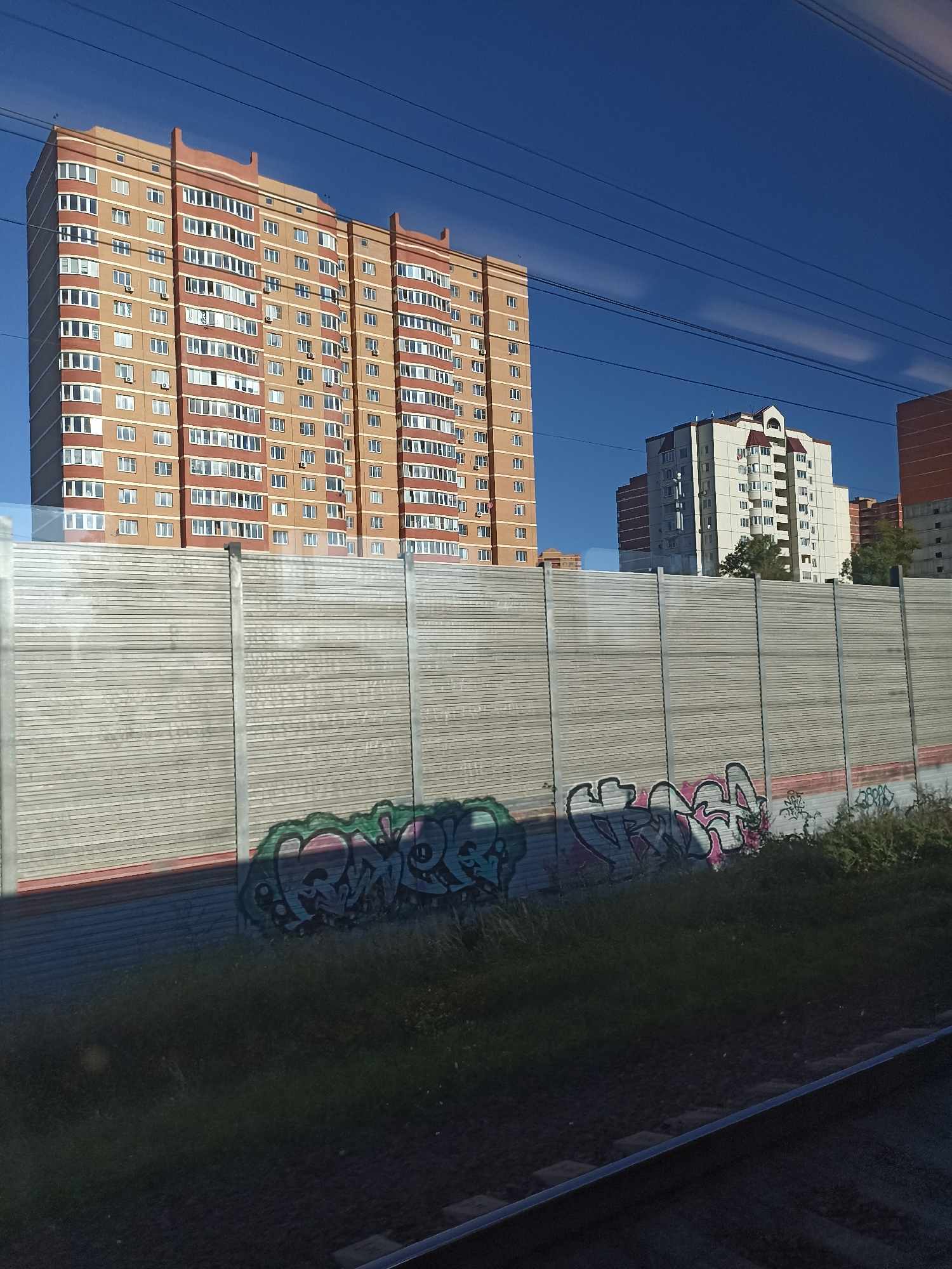 A long, tan and red apartment building with many windows is visible under a clear blue sky. In front of the building, a tall, grey, corrugated metal fence runs horizontally across the frame. Graffiti, in shades of green, pink, and black, is spray-painted onto the lower section of the fence. To the right of the main building, a lighter colored apartment building with a red roof and a peaked architectural feature is visible. More buildings, possibly under construction or with similar designs, are partially visible to the far right and behind the fence. Power lines stretch across the top of the image. The bottom of the image shows grass and what appears to be a section of train tracks, indicating the vantage point is likely from a moving train. The scene is set in the city of Shcherbinka, Russia.