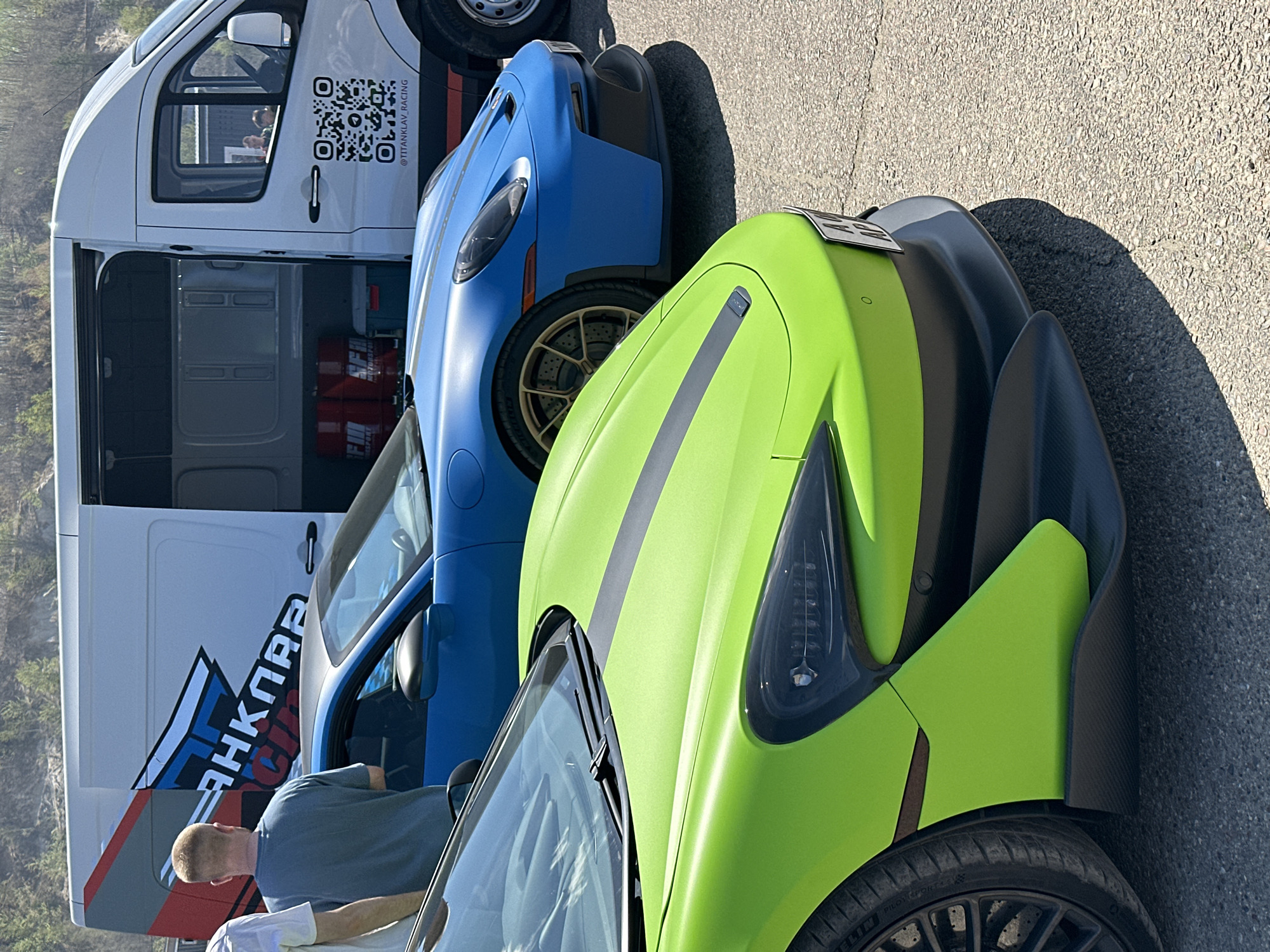 The image shows a white van parked next to a blue sports car and a lime green sports car. The van has a logo of "TITANKLAV RACING" and a QR code. A man in a grey shirt is standing near the van and the blue car. The cars are parked on an asphalt surface. The location, Sadovoe, Russia, is not verifiable in the image.