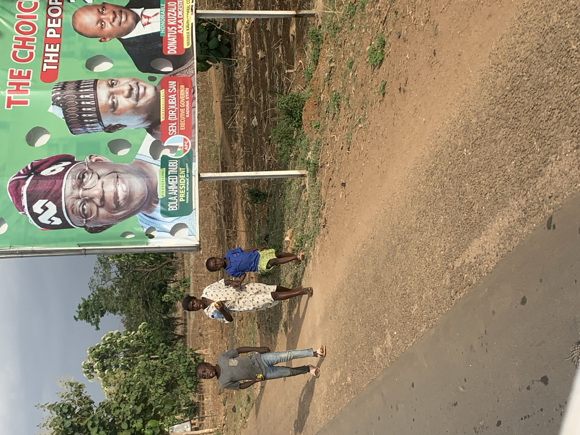 Three children stand on a road in what appears to be a rural area of Maigizo, Nigeria. A boy in a gray shirt and jeans stands closest to the camera, facing forward. A woman in a white patterned dress stands next to him. A younger child wearing a blue shirt and yellow shorts lies on the ground. Next to the children is a large political campaign sign with three portraits: Bola Ahmed Tinubu, Sen. (Dr.) Uba Sani, and Donatus Kuzalio. The sign reads "The Choice: The People." Trees and vegetation are visible in the background. The road surface is asphalt.