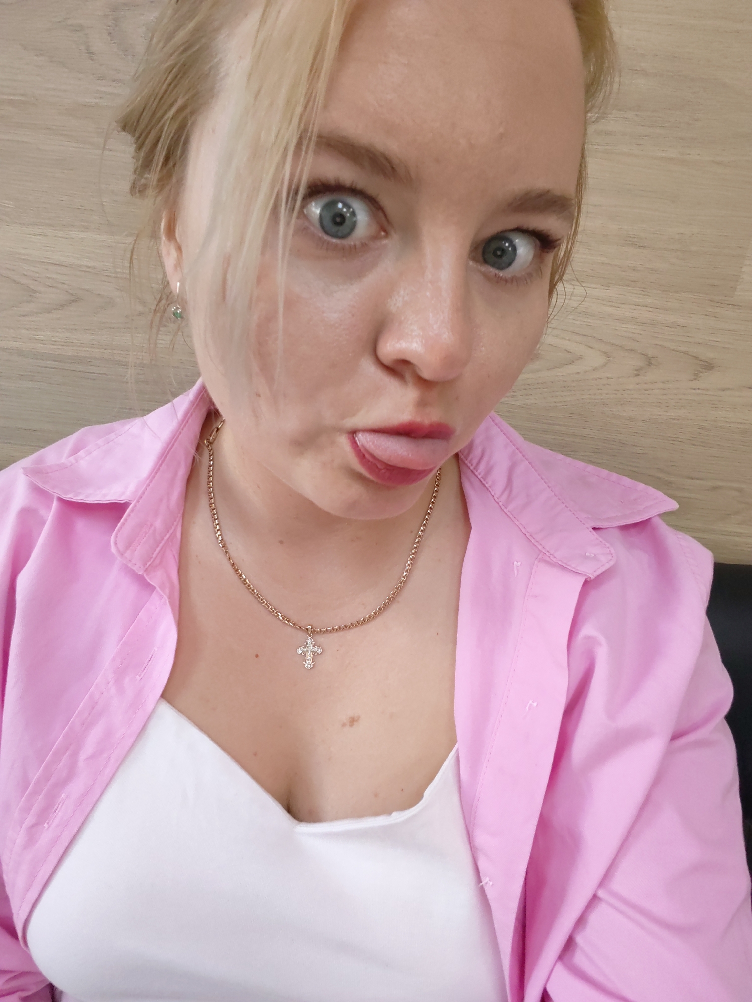 A woman with light blonde hair pulled back except for some strands near her face, is posing for a selfie. She is wearing a pink button-down shirt over a white top, and a gold necklace with a cross pendant. She is sticking her tongue out and widening her eyes, creating a playful expression. The background appears to be a light-colored wooden wall. There is no information to specify this image was taken in Izhevsk, Russia.