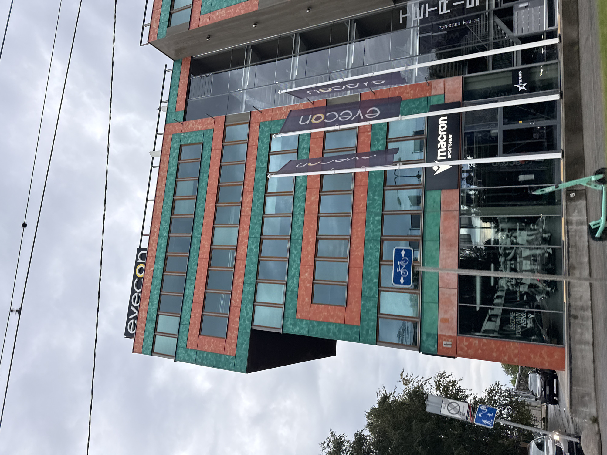 The image shows the exterior of a modern, multi-story building in Tallinn, Estonia. The building has a distinctive architectural style with alternating panels of reddish-orange and teal-green. The facade is punctuated by rows of windows. A prominent sign with the "evecon" logo is visible at the top of the building. Below the upper floors, the building features a glass-fronted section that appears to house commercial establishments. One of these is a "Macron Sports Hub" with a large window displaying athletic imagery and text that reads "BECOME YOUR OWN HERO." Three vertical banners with the "evecon" logo are displayed outside this ground-level section. Adjacent to the Macron Sports Hub, another sign reads "TEAMS." To the left of the building, a street is visible with parked cars and traffic signs, including one with a bicycle symbol and another indicating pedestrian access. A light-colored car is in the foreground on the left, and a dark SUV is on the street. A teal-colored electric scooter is parked on the sidewalk in front of the building. Overhead, power lines are strung across the cloudy sky. The overall impression is of a commercial or office building in an urban environment.