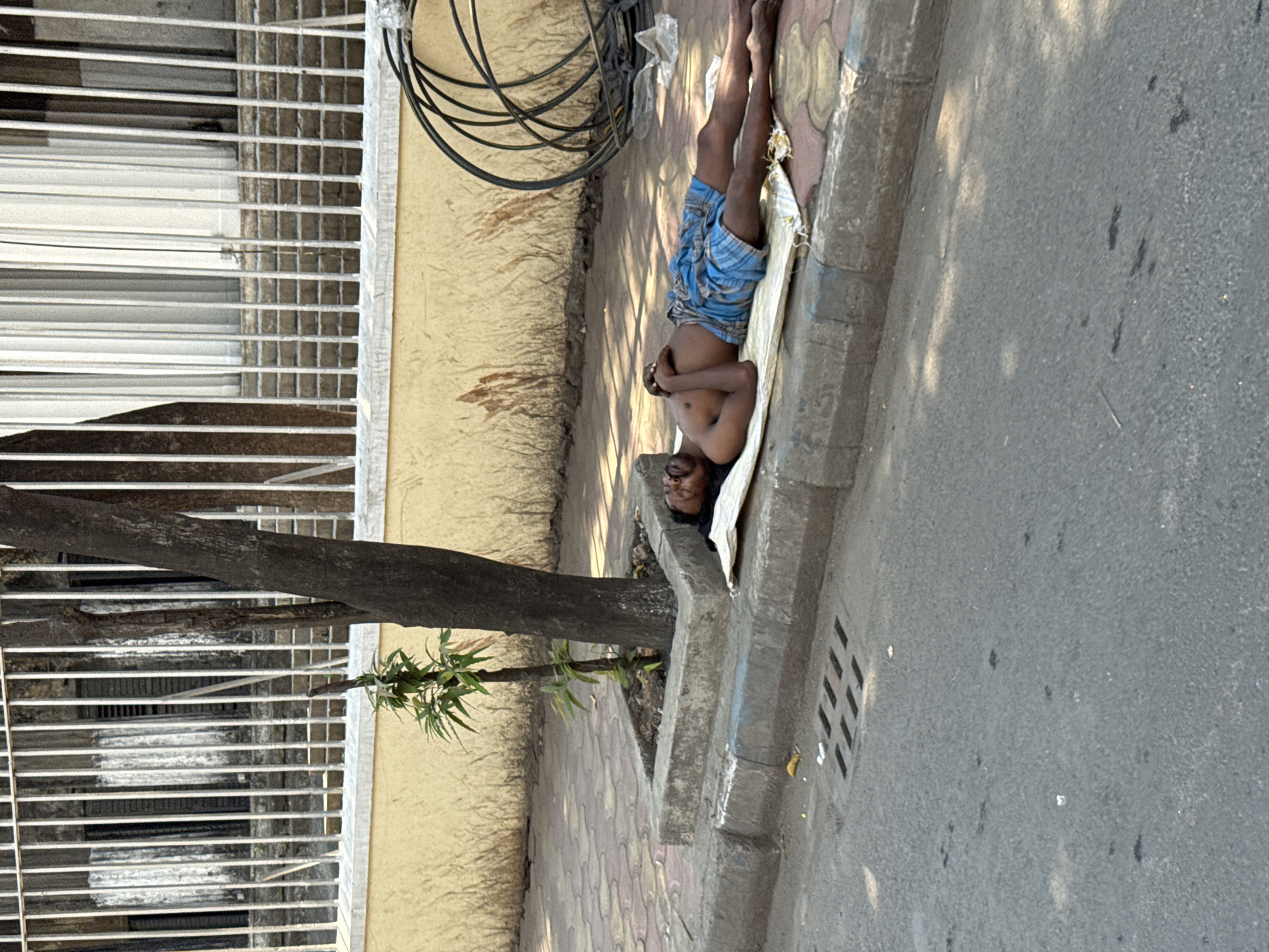 The image shows a man lying on a thin mat along the edge of a sidewalk in Kolkata, India. He is wearing blue and white checkered shorts, shirtless, and appears to be resting. He has dark hair and is wearing glasses. He is next to a building with a wall that shows signs of wear and discoloration. There is a black coiled object above him, possibly a hose or electrical cable. A small tree grows beside the curb. A section of roadway is visible adjacent to the sidewalk.
