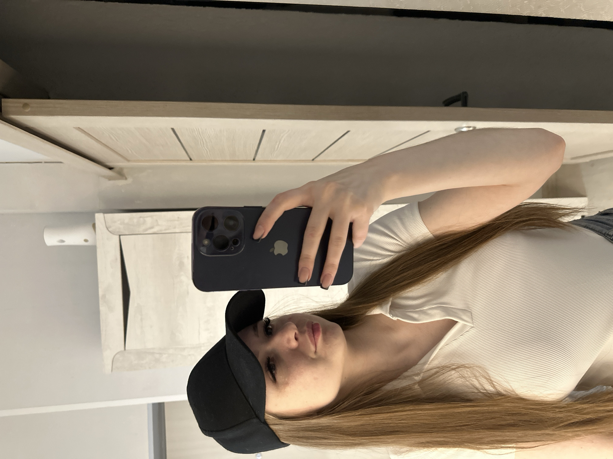 A woman wearing a black baseball cap and a white ribbed shirt is taking a selfie with a dark blue iPhone. The background shows a white cabinet, a beige surface, and a black object possibly a TV or monitor. Location: Unknown, Unknown.