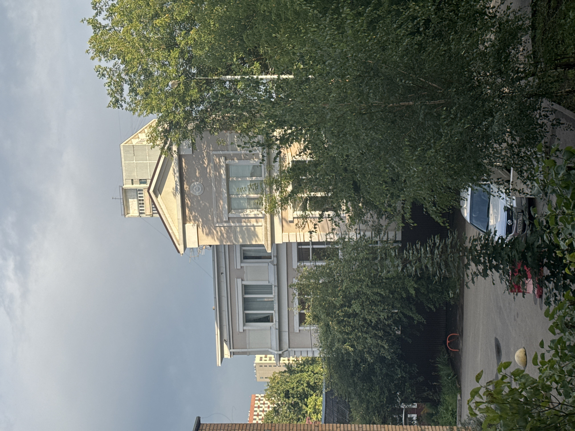 A cloudy sky looms over a residential area in Shcherbinka, Russia. In the foreground, lush green trees obscure a parking lot where a silver car is parked. Behind the trees, a light-colored, multi-story building with a triangular facade is visible. To the left of this building, another building with red and white brickwork is partially visible. In the background, taller apartment buildings rise into the sky. A dark fence and some foliage can be seen near the base of the buildings.