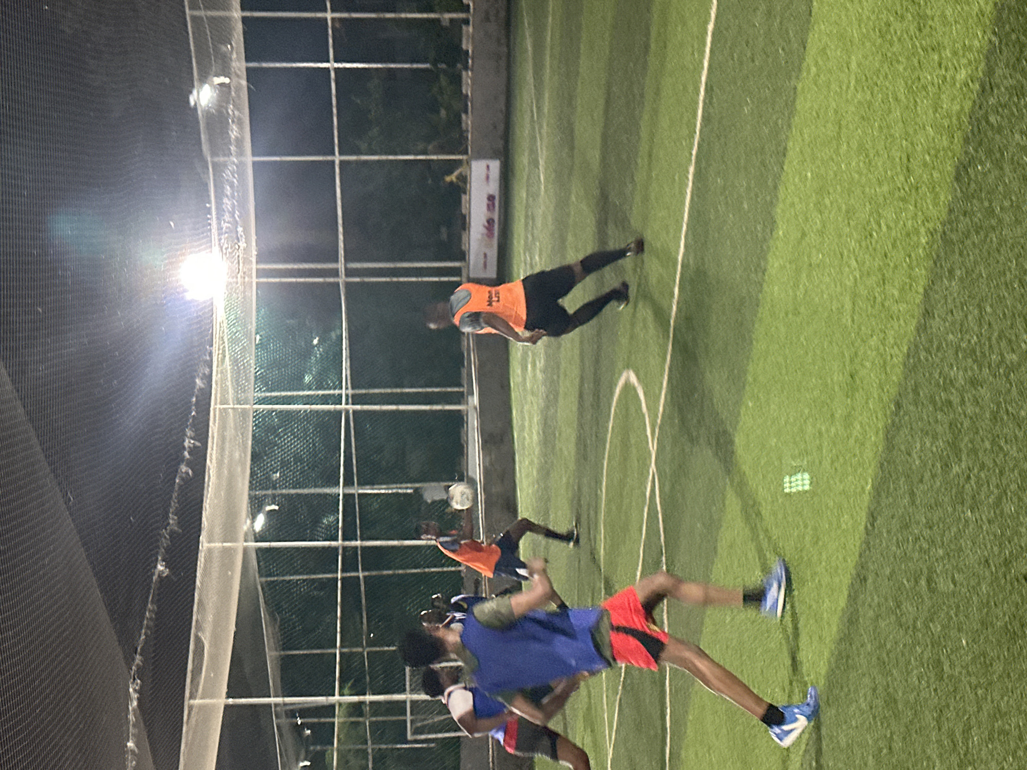 In the city of Abuja, Nigeria, a group of young men are engaged in a soccer game on an artificial turf field at night. The field is enclosed by a net and metal fencing, with bright overhead lights illuminating the playing area. Several players are visible, some wearing athletic vests over their shirts. One player in an orange vest is in mid-action, about to kick a soccer ball. Another player in a blue vest is running towards the left side of the frame, while a third player in red shorts and a blue vest is also in motion. The game appears to be a casual match, with players focused on the ball.